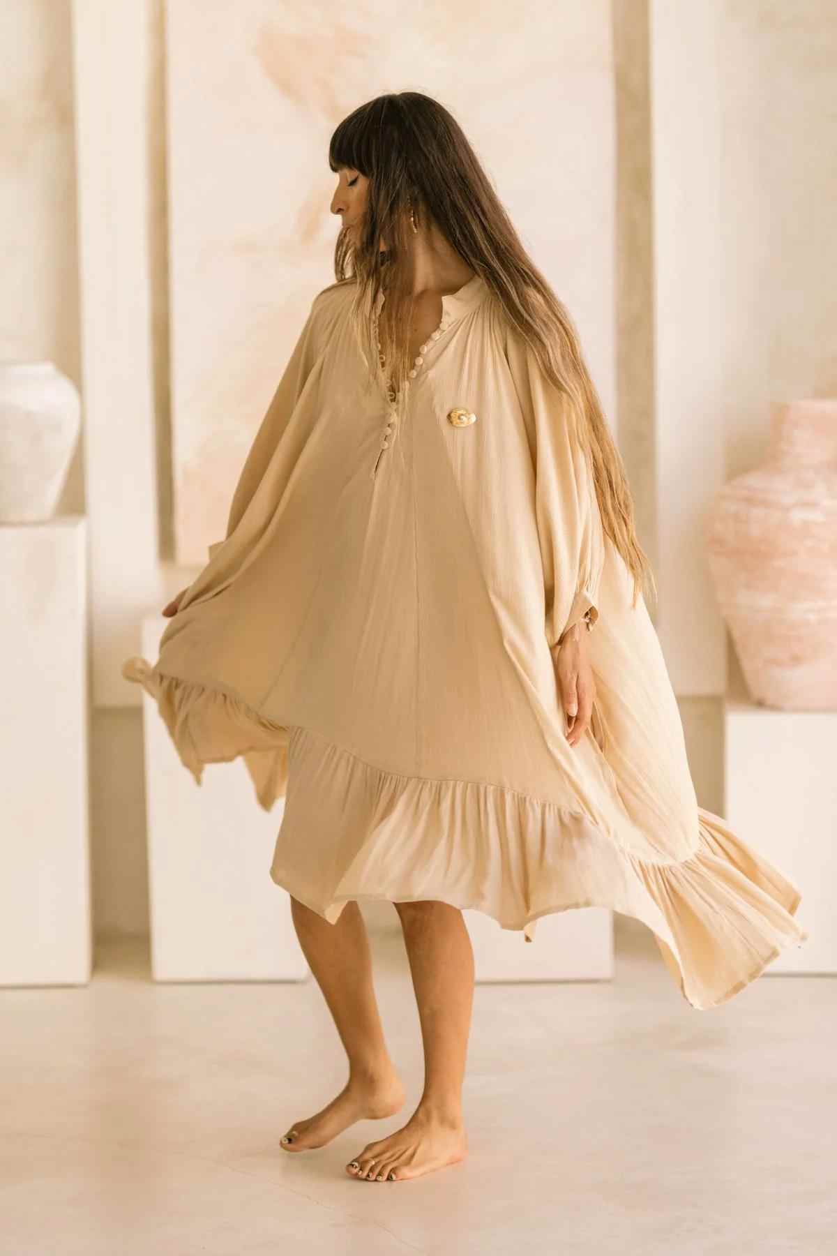 A barefoot woman with long hair is wearing a loose-fitting, beige dress indoors on a light-colored floor. The neutral-toned decor sets the backdrop as she gracefully holds her Bamboo Rayon FRIDA Gown Short by Myrah Penaloza, a true testament to handmade garments, while looking to the side.