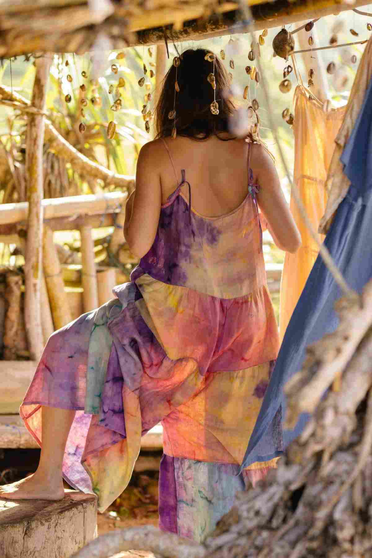 A person with long hair is seated on a wooden platform, wearing the vibrant La Mexicana Playsuit by Myrah Penaloza, crafted from 100% linen and made to order. Seashells hang above, enhancing the bohemian atmosphere as sunlight filters through the trees in the background.