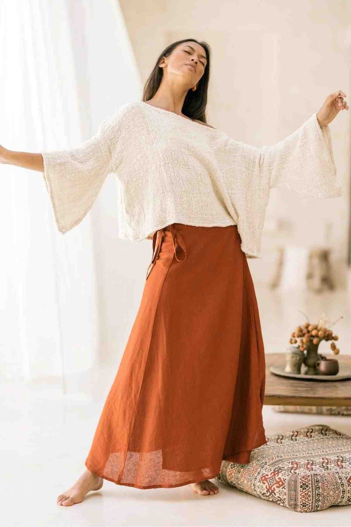 A woman is standing with her arms slightly raised, dressed in the Spirit Top and Shanti Wrap Skirt Set from Myrah Penaloza. She appears to be in a bright, airy room with soft natural light. A low table with a fruit bowl and a patterned cushion made from Hand-Woven Tummangal Cotton is visible in the background.