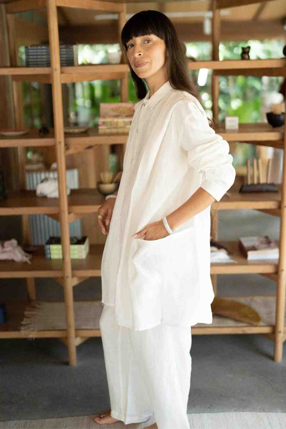 A person with long dark hair stands indoors in front of a wooden bookshelf, wearing an oversized white button-down shirt, white pants, and the Tender Jacket (100% Linen) by Myrah Penaloza. They have one hand in their pocket and are smiling slightly. The room appears to be well-lit and filled with books and decor items, reflecting an eclectic fashion style.