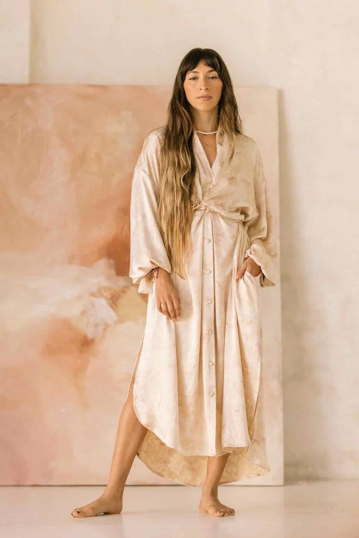 A woman with long, wavy hair stands barefoot indoors, wearing a beige Silk Inanna Wrap Gown by Myrah Penaloza made of botanically hand-dyed silk. She stands in front of a large, abstract pink and beige painting, looking directly at the camera with a calm expression.