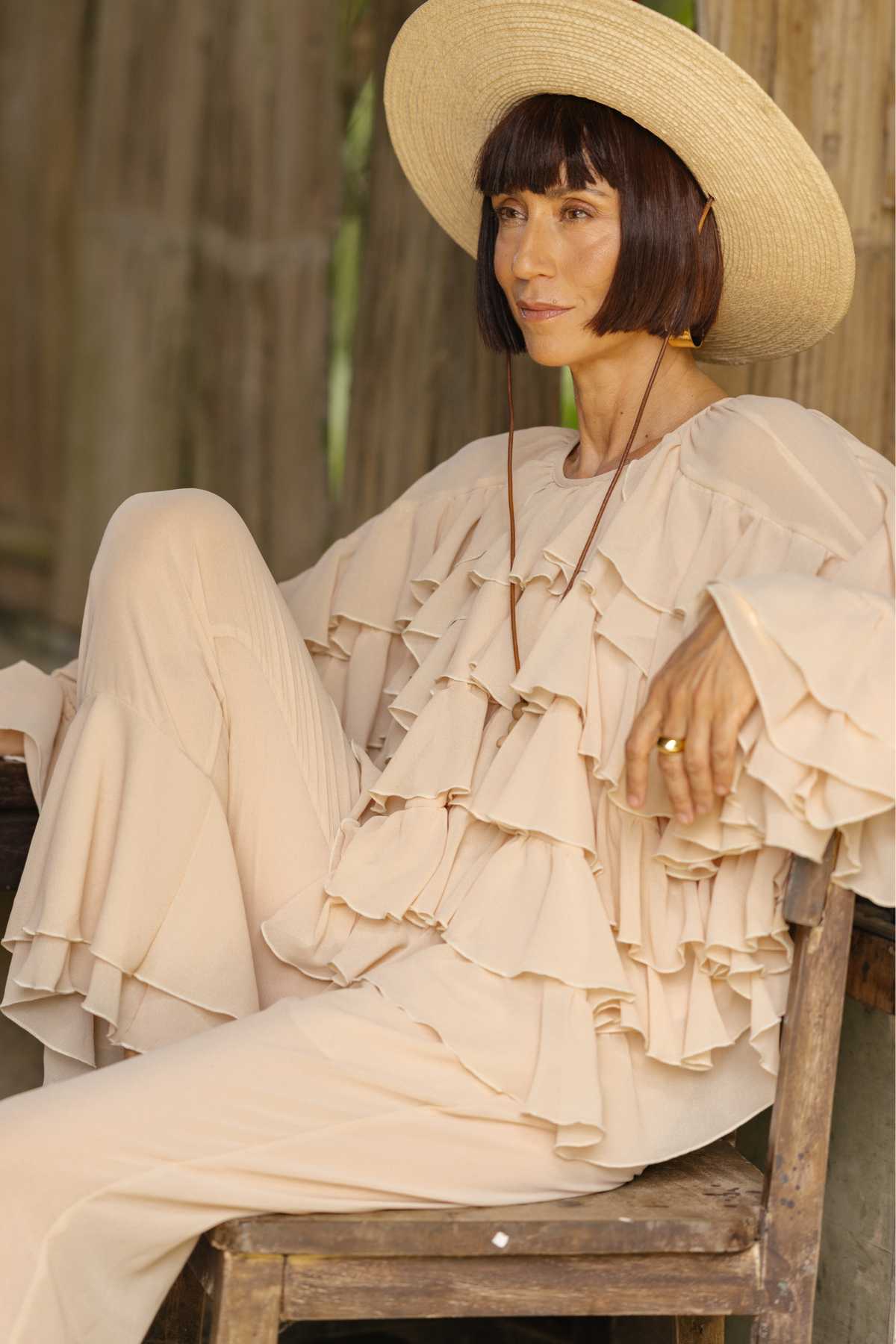 A person with short, dark hair is seated on a wooden chair, wearing a wide-brimmed straw hat and an outfit with multiple ruffles in a light, neutral color. Their attire appears to be the nature-inspired couture "Anusara Bloom - Made To Order" by Myrah Penaloza, crafted in limited edition chiffon sheer silk. The setting is outdoors with blurred wooden structures in the background.