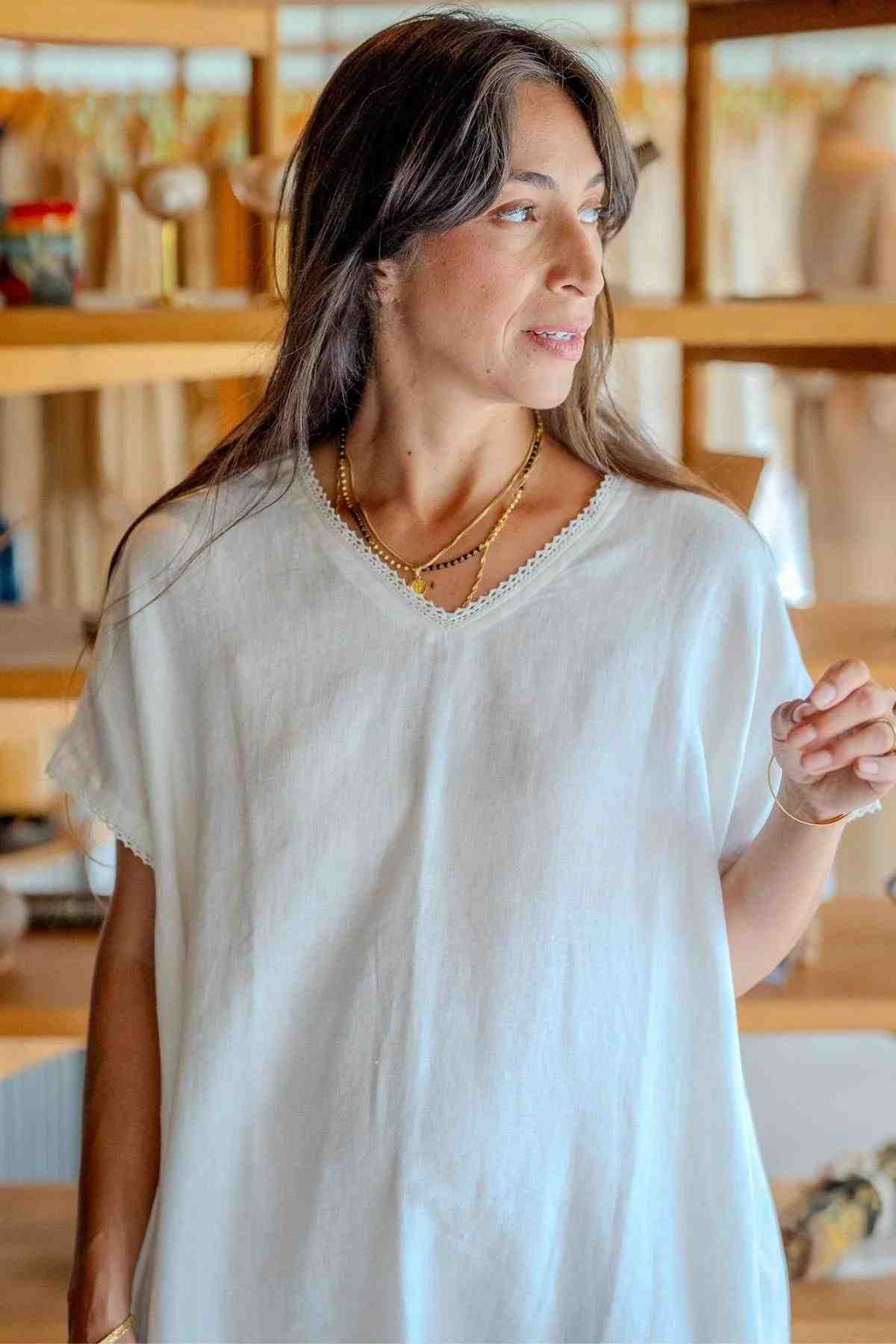 A woman with long dark hair stands indoors, wearing the Ruby Lace Tunic from Myrah Penaloza, featuring a delicate V neckline. She has multiple gold necklaces layered around her neck and is looking off to the side. Behind her, there are shelves with various items.