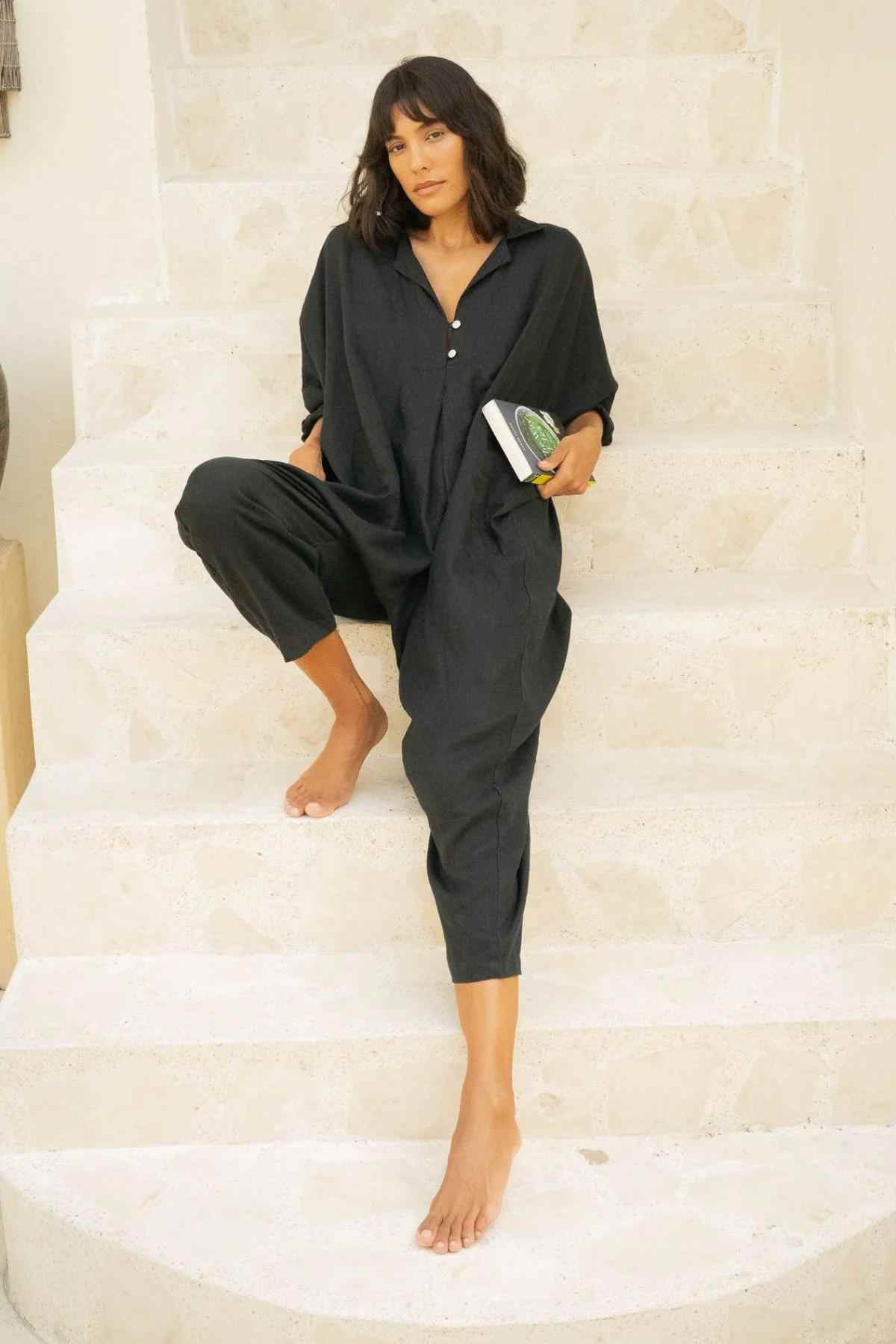 A woman with shoulder-length hair, dressed in a loose Linen Sat Torri Playsuit from Myrah Penaloza featuring wide dolman sleeves, sits on white marble steps. She holds a book in her right hand and rests her left hand on her bent knee. Her expression is relaxed and she is barefoot, exuding a retro 1980s inspired design vibe.