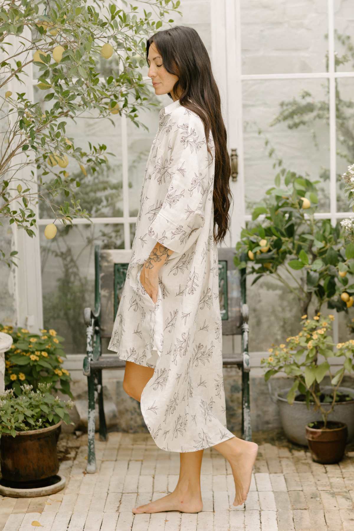 A woman with long dark hair, seen in profile, is wearing a long, flowy cream-colored Linen Suka Button Down Long Top by Myrah Penaloza. The sustainable fabric features a delicate floral pattern. She stands barefoot on a tiled floor in a greenhouse, surrounded by potted plants and citrus trees, as sunlight filters through the large windows.