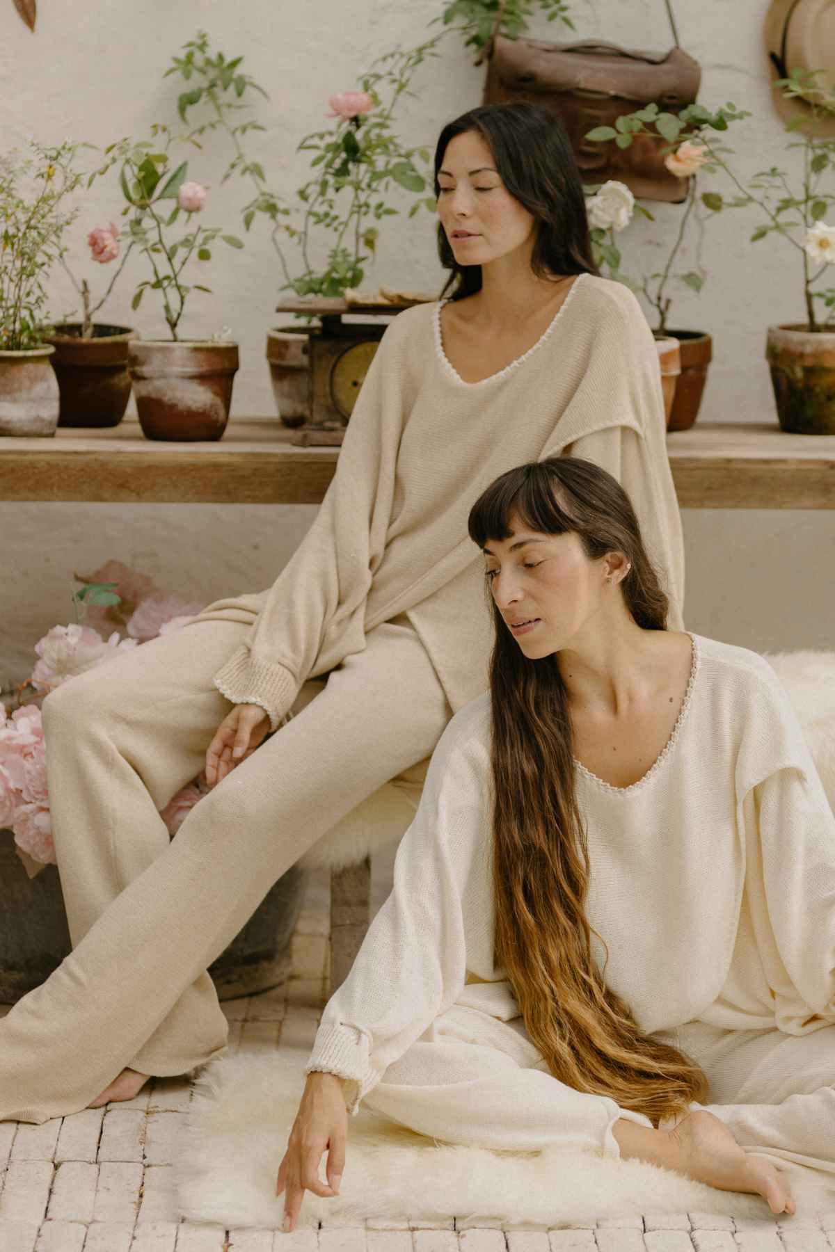 Two women dressed in Myrah Penaloza's soft, neutral-colored Honey Knit Sweaters are relaxing in a cozy setting with potted plants. One is seated on a bench with eyes closed, while the other sits on the floor, touching a white rug. The atmosphere is calm and serene.