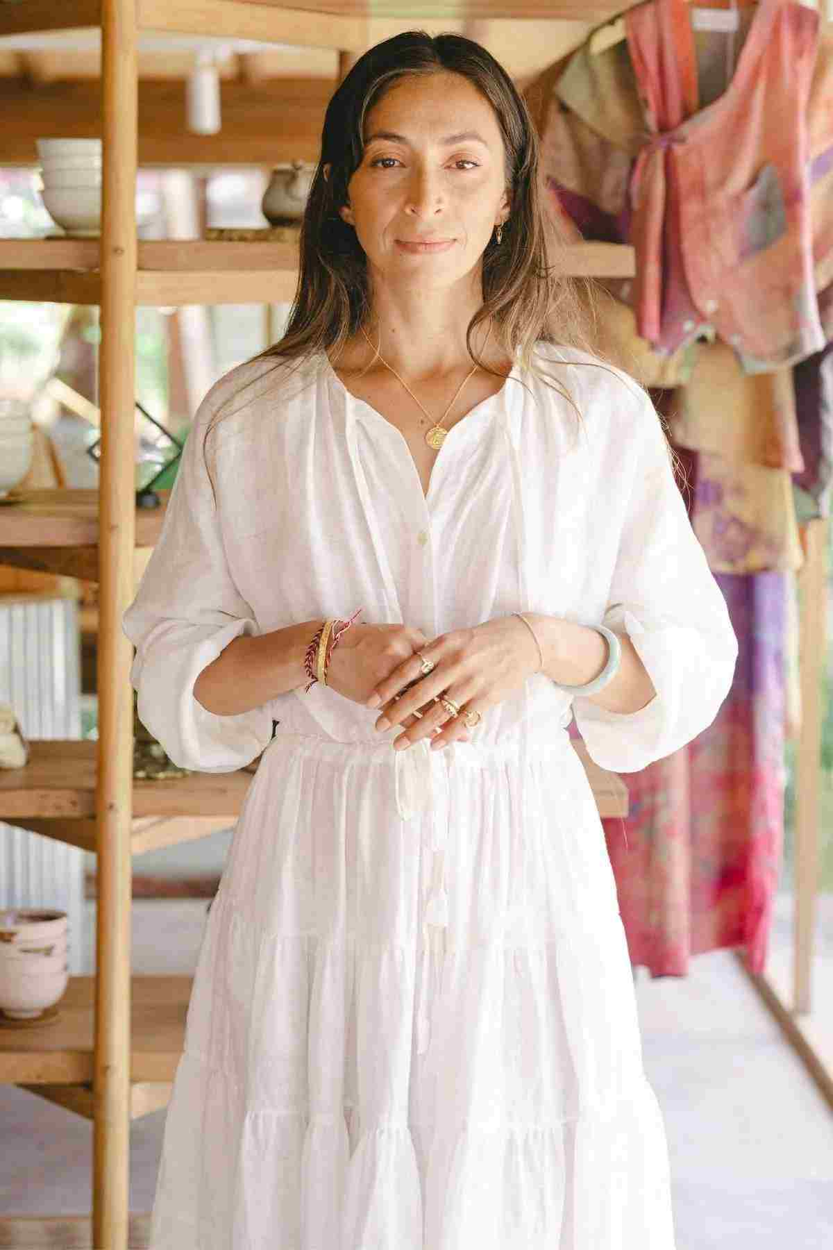 A person wearing the La Mexicana Gown by Myrah Penaloza, featuring an adjustable waist and available in multiple colors, stands inside a room with wooden shelves in the background. The shelves display pottery and vibrant textiles. The individual has long brown hair and is adorned with jewelry.