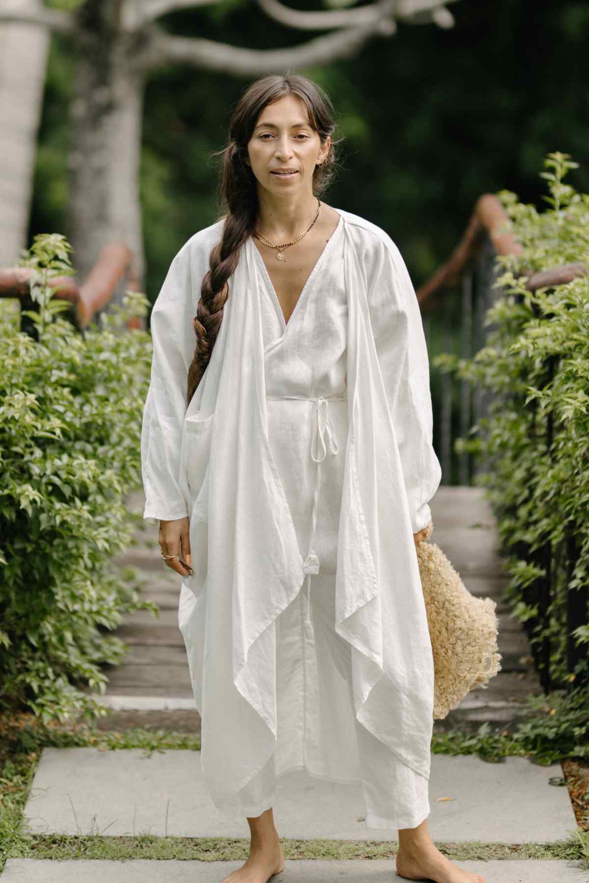 A woman with long, dark braided hair stands barefoot on a stone pathway surrounded by green foliage. She is wearing a flowing, white Dao Robe (100% Linen) from Myrah Penaloza and holds a beige, textured bag at her side. She gazes calmly at the camera with a serene expression, embodying natural colors.