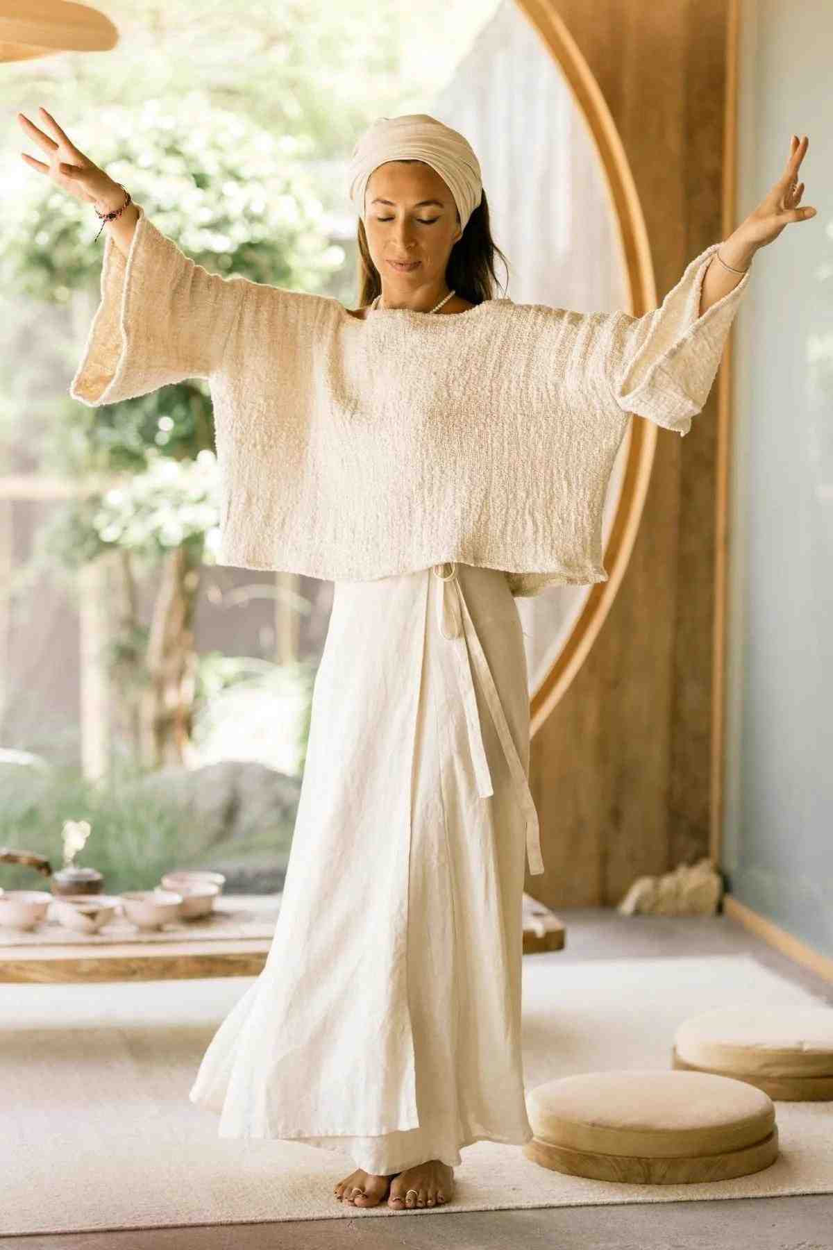 A woman stands barefoot indoors with her arms raised, wearing a loose, light-colored top and a long, flowing Myrah Penaloza Shanti Wrap Skirt made of 100% linen. She has a serene expression with her eyes closed and is wearing a headscarf. The room has a peaceful ambiance with wooden decor and soft natural light.