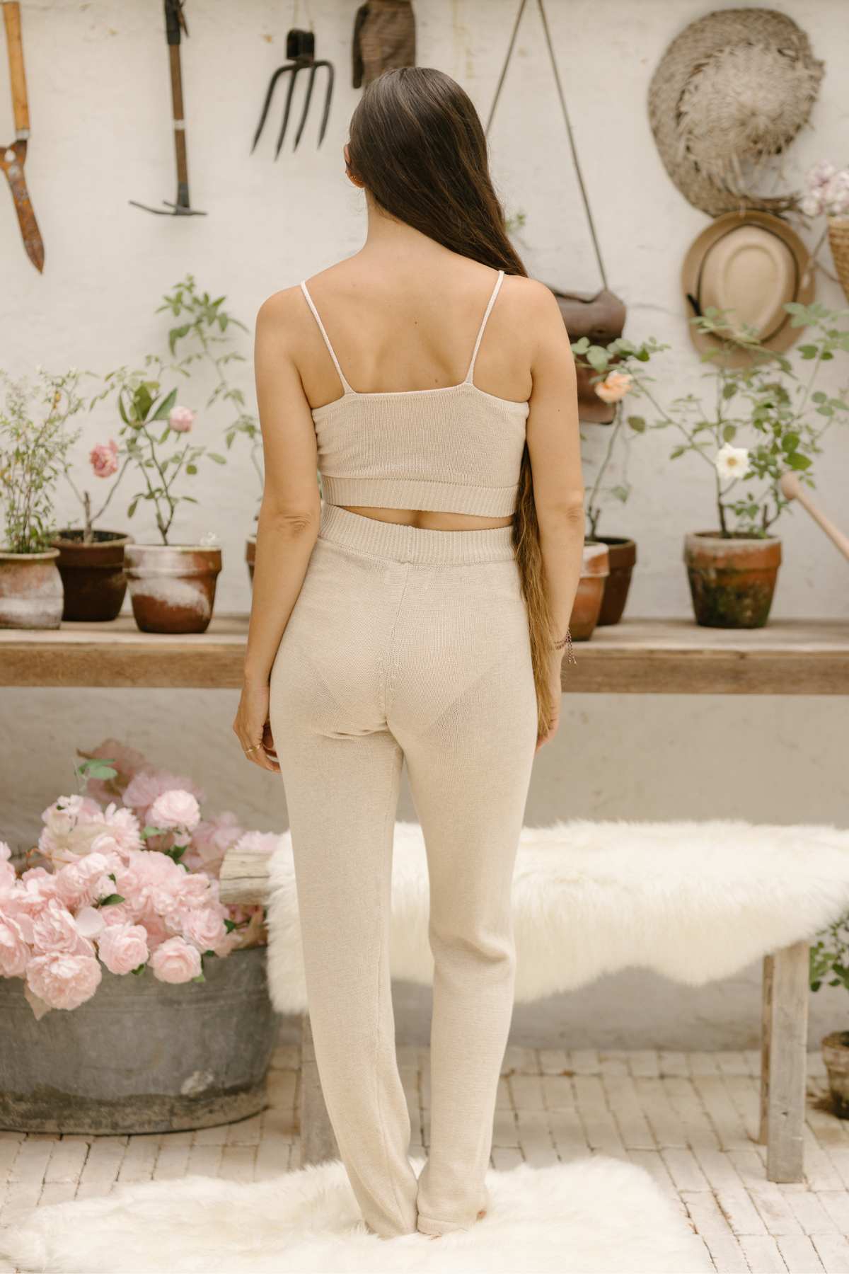 A person with long hair stands facing away, dressed in the Honey Knit Sets by Myrah Penaloza. They're in a garden setting featuring potted plants on a wooden table, standing on a soft white rug, with gardening tools hanging on the wall.