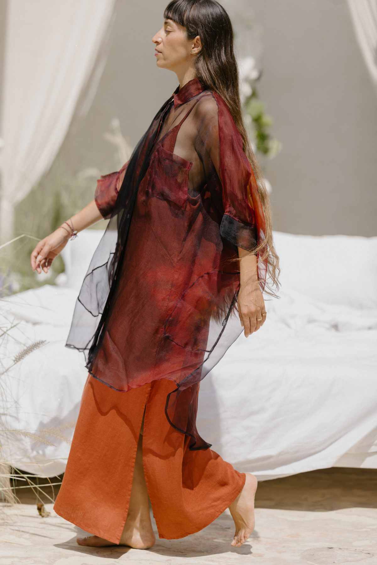 A woman with long hair walks barefoot next to a white bed, wearing an airy, sheer dark red Rainbeau Chiffon Silk Suka Top from Myrah Penaloza over an orange skirt with a side slit. The oversized fit offers versatile styling options. Off-white drapes and greenery in the background create a serene, natural setting.