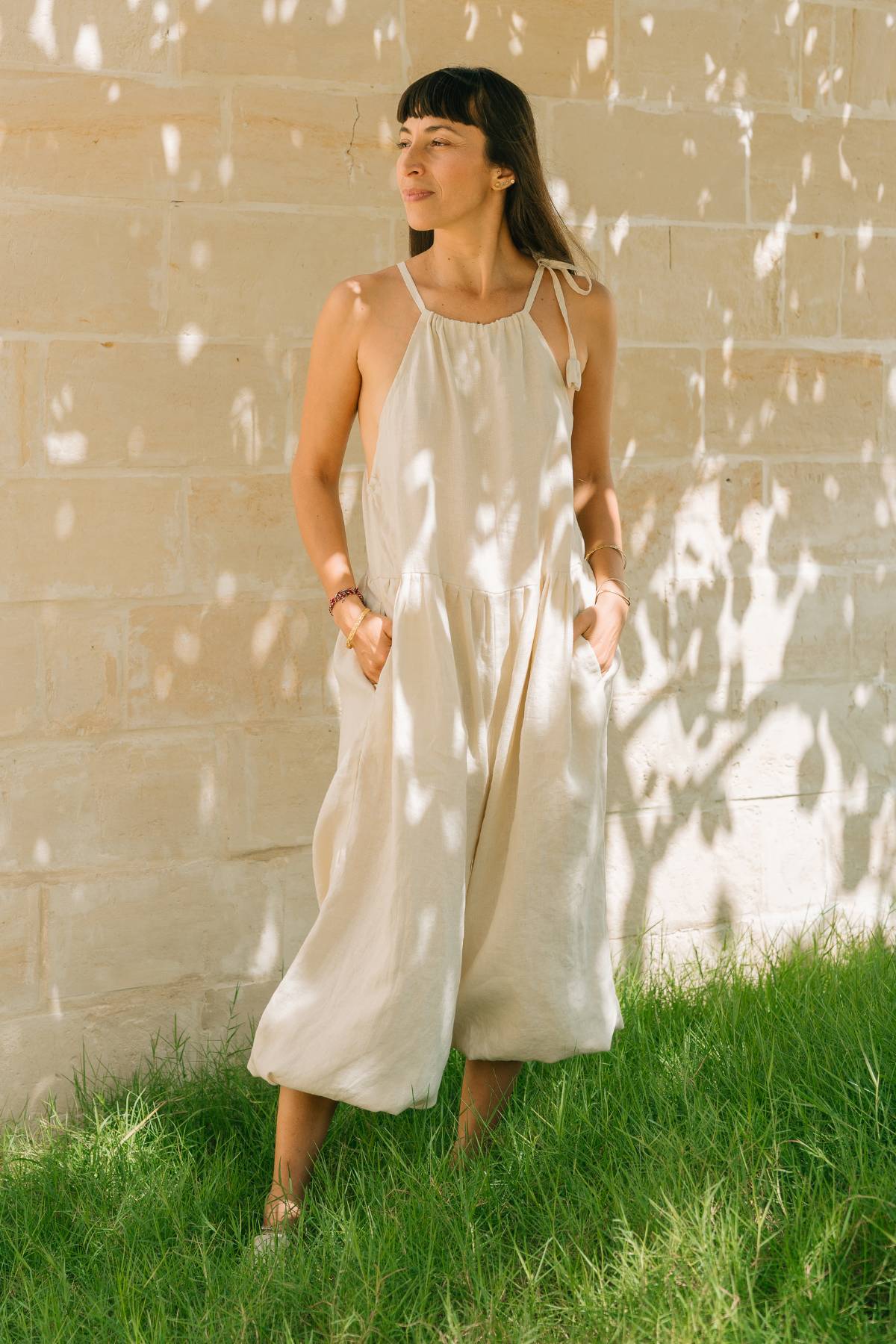 Front view of the Monaya Playsuit by Myrah Penaloza in Moonlight stone washed linen, hands in pockets, standing on grass against a stone wall. Oversized full length silhouette, handcrafted in Bali.