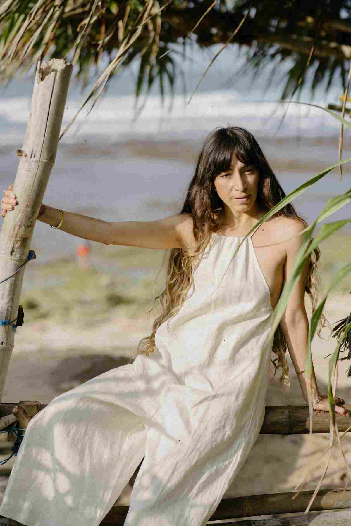 A woman with long, wavy hair sits on a wooden structure by the beach, wearing the luxurious Linen Vata Playsuit from Myrah Penaloza. She holds onto a vertical wooden pole while green plants and ocean waves can be seen in the background.