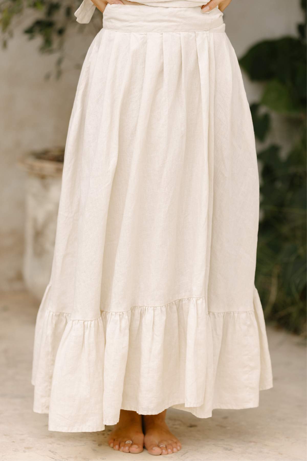 A person stands barefoot on a light flax surface, wearing a long, flowing Viva Skirt in cream-colored, stone-washed linen by Myrah Penaloza. The skirt showcases a ruffled hem and pleated design, with lush green foliage softly blurred in the background.