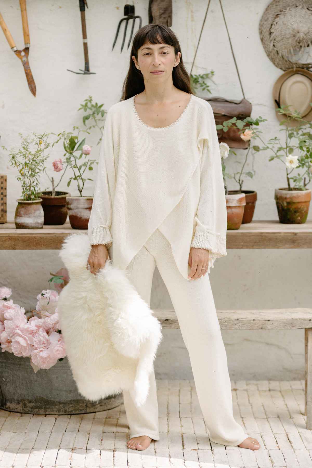 A woman stands in front of a garden potting bench filled with plants and flowers. She is wearing a Honey Knit Sweater by Myrah Penaloza and loose pants, holding a fluffy white throw. Gardening tools and a straw hat hang on the wall behind her. The scene is bright and serene.