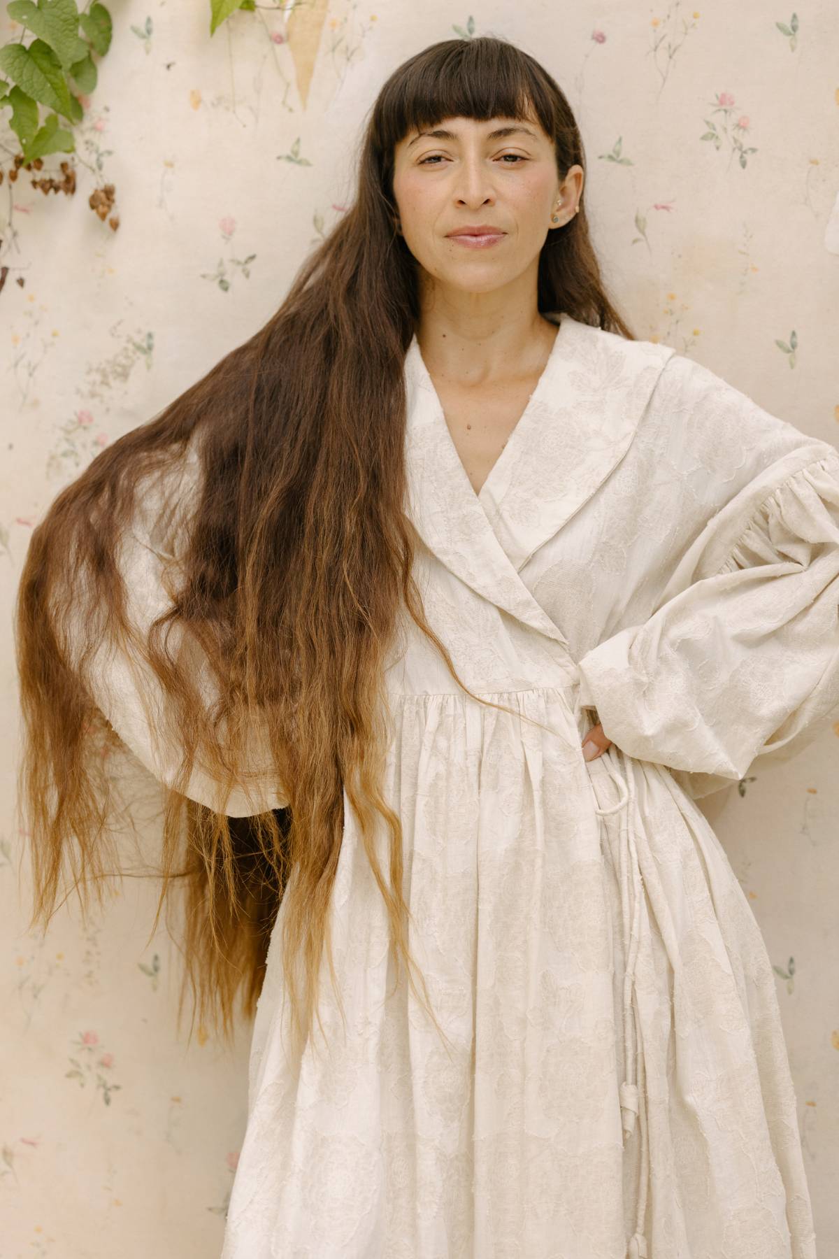 A person with long brown hair stands against a backdrop adorned with floral patterns. They are wearing the Blooming Magdalena Dress by Myrah Penaloza, featuring light-colored, long sleeves. With a relaxed expression and one hand placed on their hip, they exude an effortless elegance.