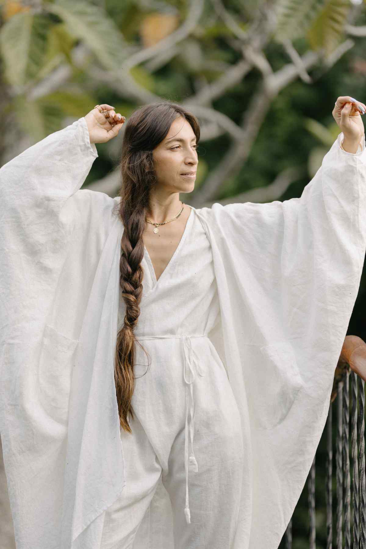 A person with long, braided hair stands outdoors, enveloped in the serene, natural colors of a loose-fitting Dao Robe made of 100% linen by Myrah Penaloza. The stone washed linen robe features wide sleeves and flows gracefully as they raise their arms and close their eyes, appearing completely relaxed and in harmony with the verdant backdrop.