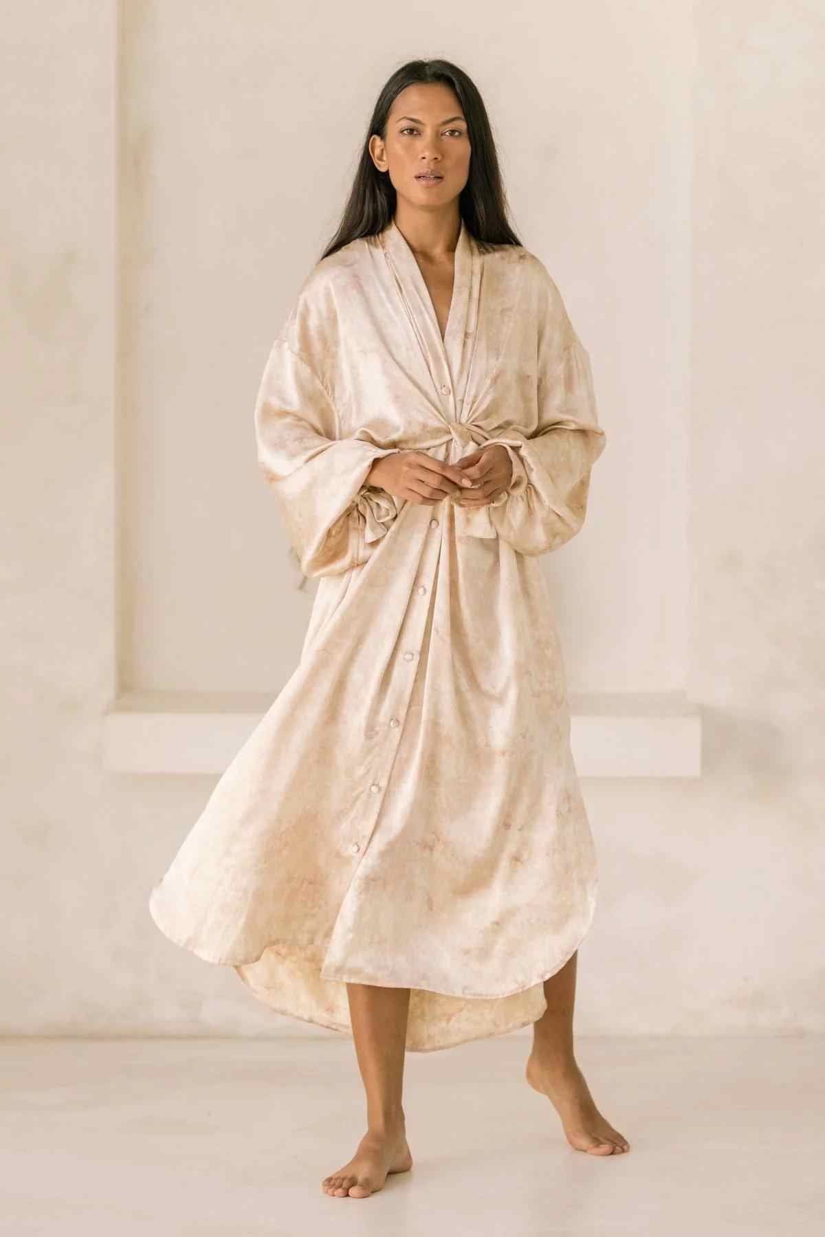 A woman stands barefoot against a plain backdrop, wearing the Silk Inanna Wrap Gown by Myrah Penaloza, a long, button-up robe made of botanically hand-dyed silk with wide sleeves. Her hands are loosely held together at her waist. The gown flows elegantly around her legs, showcasing the fine craftsmanship of artisan tailors.