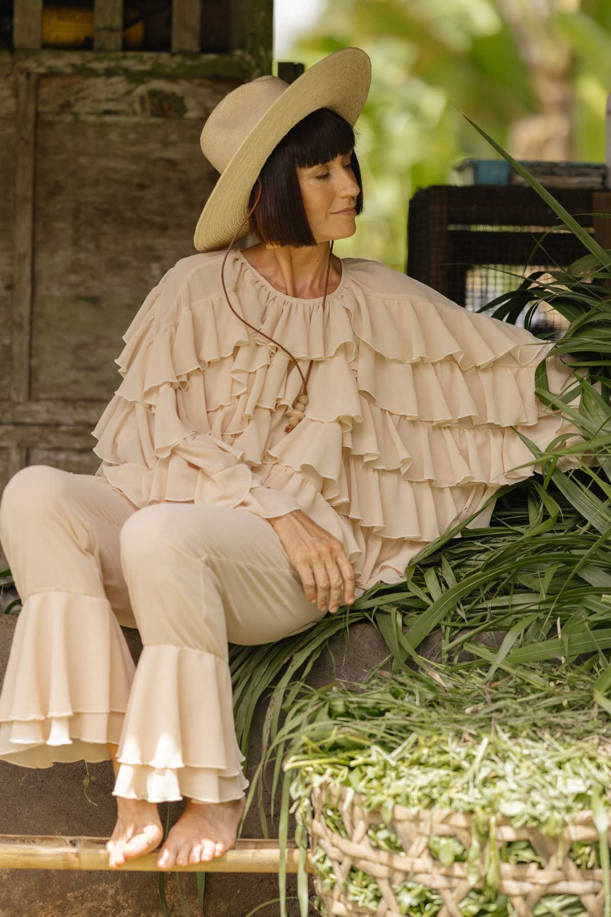 A woman dressed in the "Anusara Bloom - Made To Order" outfit by Myrah Penaloza sits outdoors on a stone ledge surrounded by lush greenery. With a bob haircut and a wide-brimmed straw hat gracing her head, she appears relaxed, gently touching the plants with one hand and looking down with a serene expression.