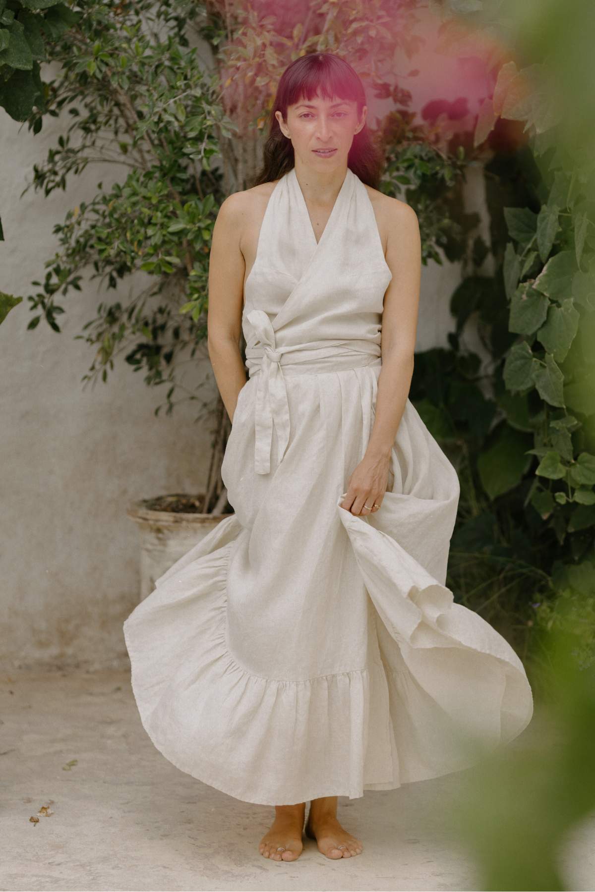 A woman stands barefoot in a cream-colored, halter-neck dress crafted from stone-washed linen, featuring a tied waistband. Surrounded by lush green plants and set against a light-colored wall, she gazes forward with a soft expression as the dress flows gracefully, reminiscent of Myrah Penaloza's Viva Skirt.