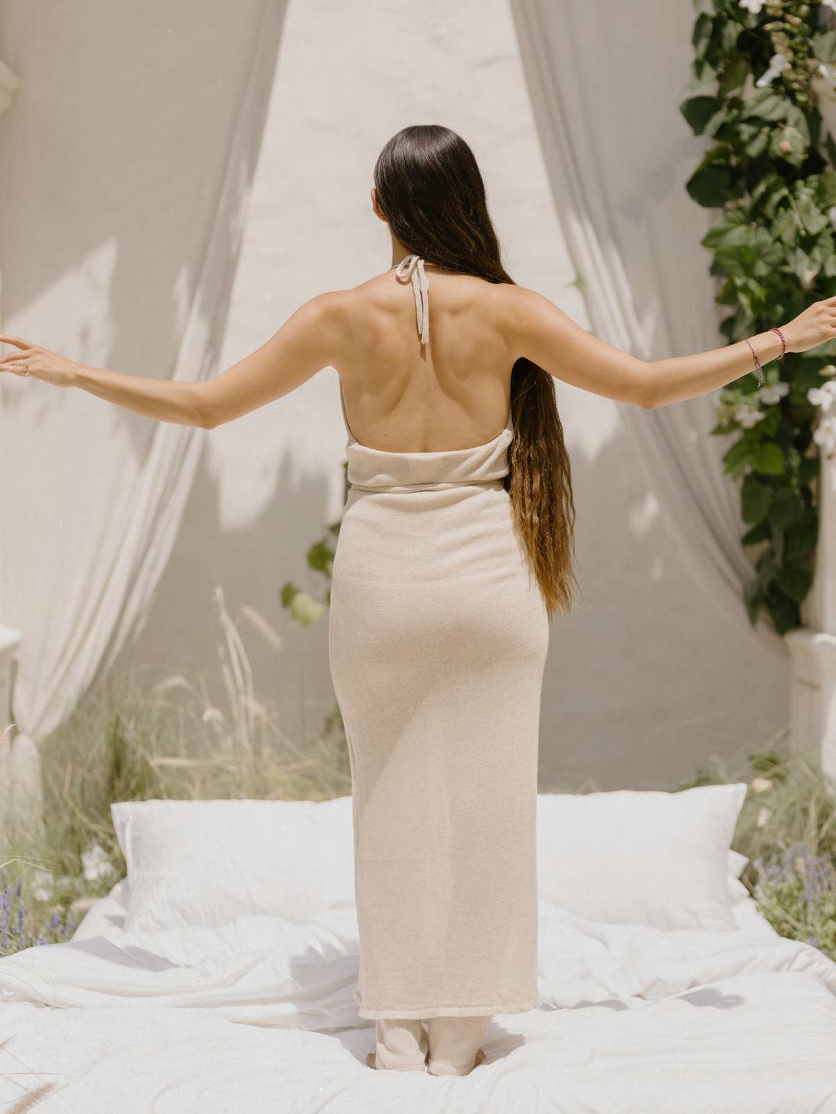 A woman, with long hair cascading down her back, stands on an outdoor bed facing away. She is wearing the Honey Cotton Knit Wrap Dress by Myrah Penaloza, its strapless beige design beautifully complementing her outstretched arms. The scene is framed by flowing white curtains and lush green foliage, creating a peaceful and natural ambiance.