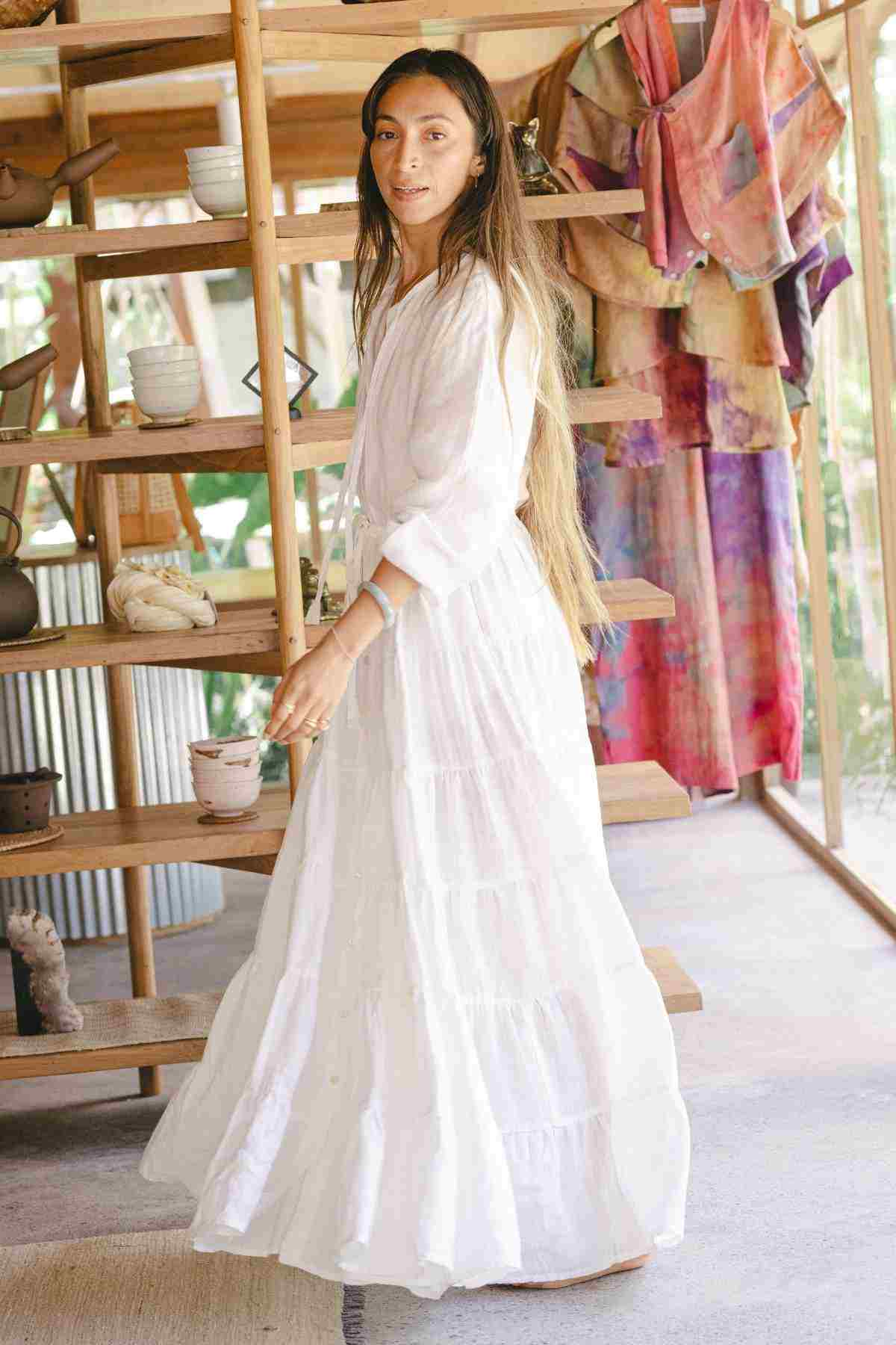 A woman wearing the La Mexicana Gown from Myrah Penaloza stands in a room with wooden shelves filled with pottery and vibrant fabrics. Sunlight streams through a large window, highlighting her long hair and the airy atmosphere. The gown, available in multiple colors, features an adjustable waist that adds elegance to the serene scene.