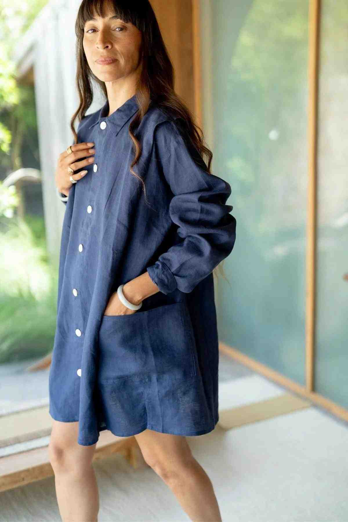 A person with long hair is standing indoors near a wooden and glass wall. They are wearing a loose-fitting, button-up Tender Jacket in deep royal blue from Myrah Penaloza, with their hands in the pockets. Soft natural light illuminates the space, highlighting their eclectic fashion sense.