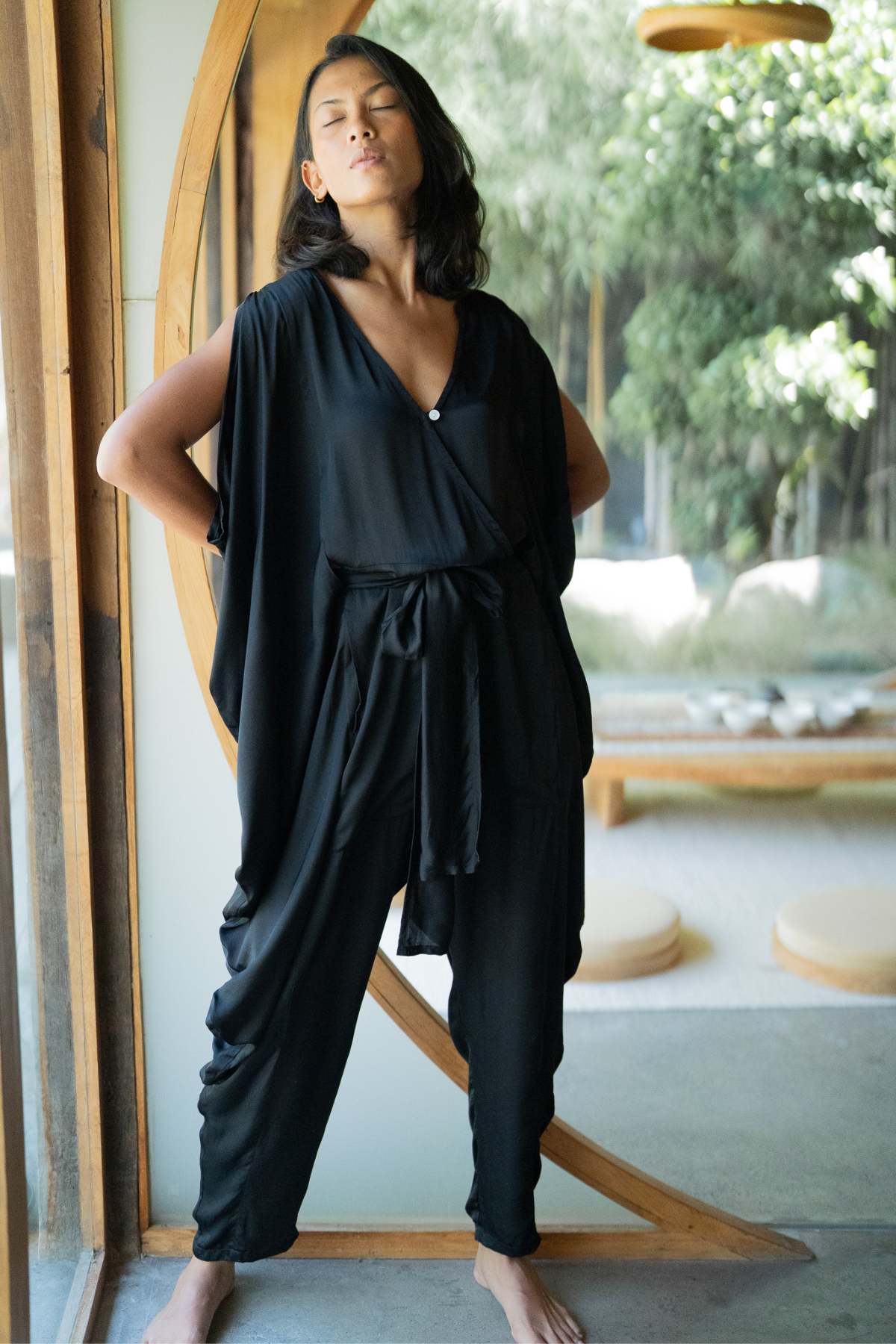 A person stands indoors, leaning against a circular wooden frame, wearing the Kuan Yin Playsuit from Myrah Penaloza, a 100% silk, botanically dyed Turmeric Gold ensemble made to order. The playsuit features an adjustable linen waist tie. They have long hair and are barefoot, posing confidently as soft natural light filters through large windows behind them.