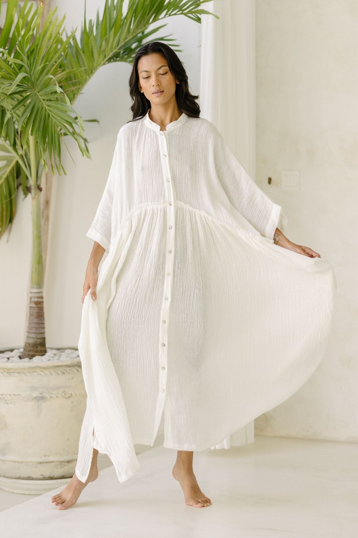 A woman wearing the flowing, long white Bona Dress (Cotton/Linen Crinkle) from Myrah Penaloza stands barefoot inside a light-filled room. Her dress features buttons down the front, and she gracefully holds out the sides, showcasing its airy fabric. A large potted plant is visible in the background.