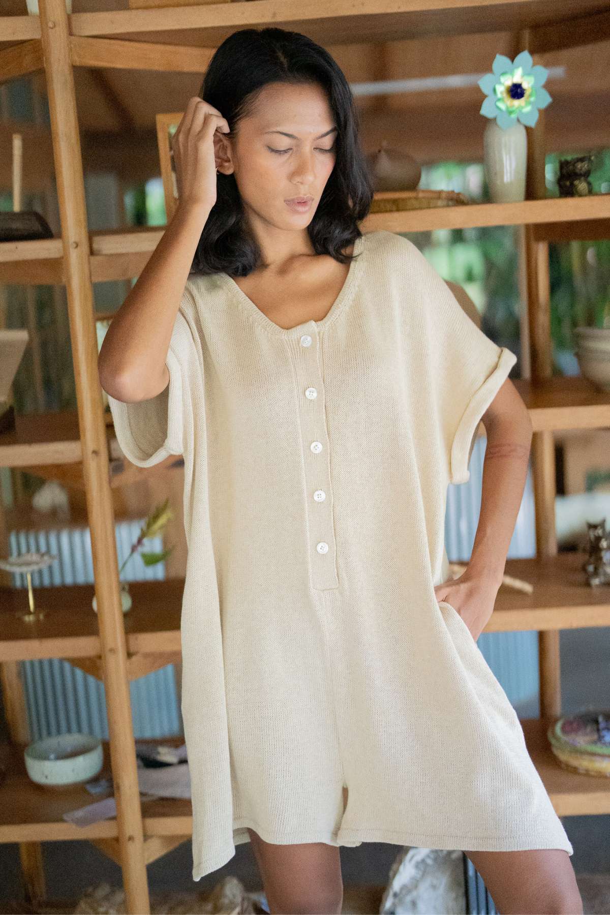 A person stands in a beige, short-sleeved romper that brings to mind the Henley Knitted Playsuit - BF by Myrah Penaloza, featuring a button-down front. The cozy cotton knit design is displayed against wooden shelves adorned with decorative items, while the softly lit background creates a warm and inviting atmosphere.