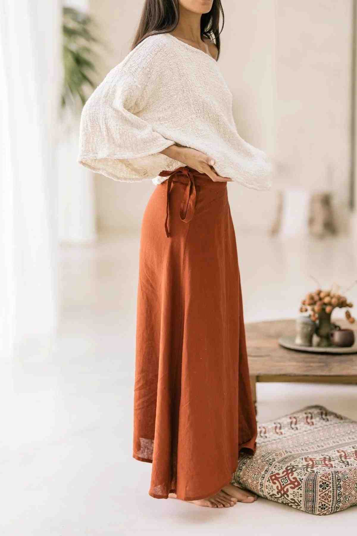 A woman stands indoors, wearing a loose, off-the-shoulder white Spirit Top and a long, flowing Shanti Wrap Skirt tied at the waist from Myrah Penaloza. She is barefoot, and the background features minimalistic decor with neutral tones, including a patterned cushion and a tray with decor items.