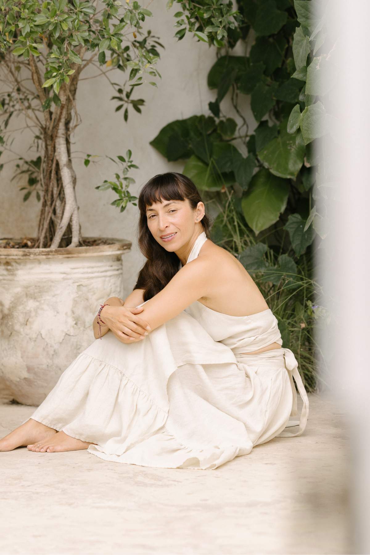 A woman in a sleeveless white dress sits barefoot on a stone floor, her arms resting on her knees as she smiles amidst lush green plants in a tranquil, natural environment. She is wearing the Viva Skirt by Myrah Penaloza, crafted from light flax for an effortlessly elegant look.