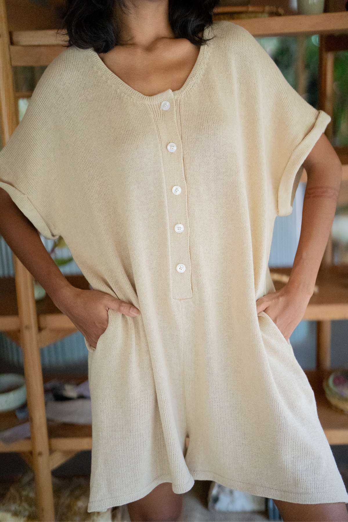 A person is photographed wearing the Henley Knitted Playsuit - BF from Myrah Penaloza, a short-sleeved, beige romper featuring button-down detailing, standing confidently with their hands in pockets. This cotton knit playsuit blends style and comfort effortlessly against the backdrop of wooden shelves, designed as an ideal breastfeeding-friendly option.