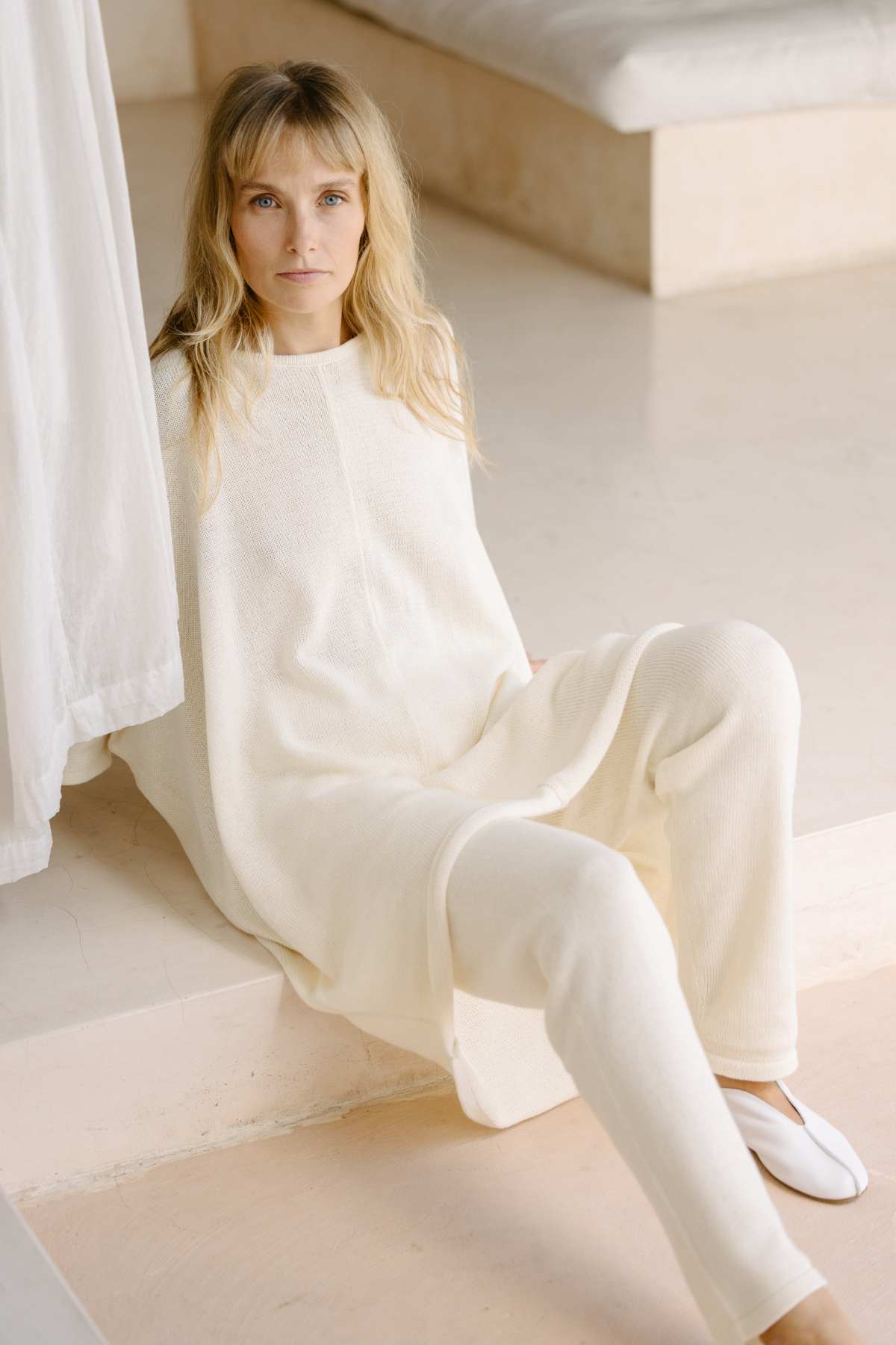 A person with long blonde hair is sitting on a step in a minimalist room. They are wearing the Butterfly Tunic Cotton Knit, a loose-fitting, cream-colored outfit from Myrah Penaloza, made from sustainable fabric, paired with white shoes. The background features neutral-toned surfaces and soft lighting, creating a calm and serene atmosphere.