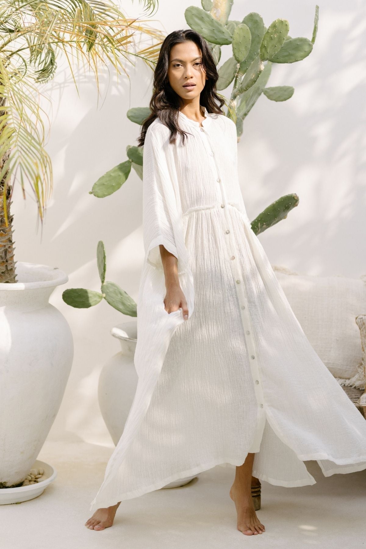 A woman stands barefoot on a sunlit patio, wearing a long, flowing Bona Dress (Cotton/Linen Crinkle) (PR) from Myrah Penaloza with buttons down the front. The elegant texture of the Cotton/Linen Crinkle fabric complements the serene and natural setting, surrounded by large potted plants such as a tall cactus and a palm—perfect for holiday season events.