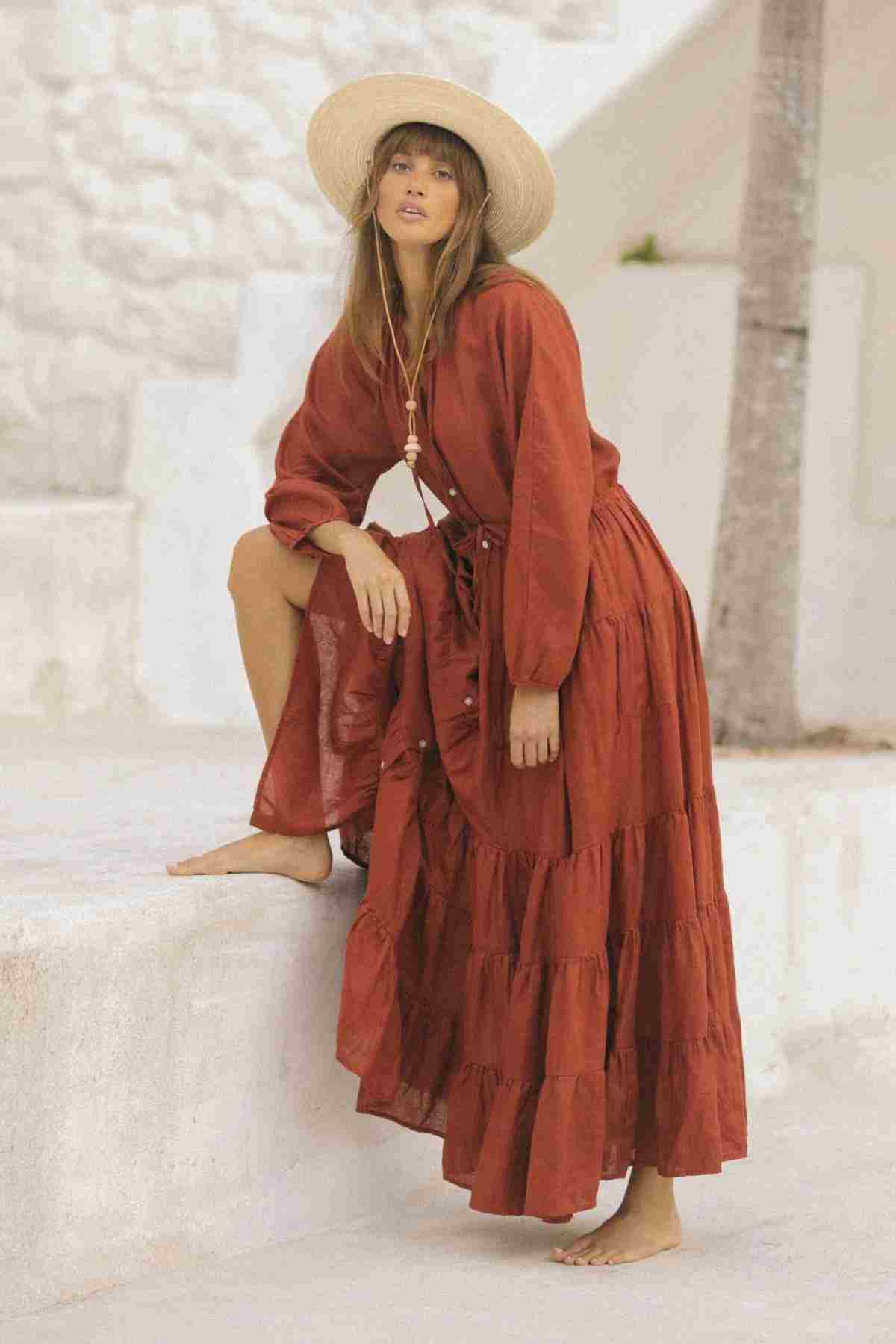 A person wearing the La Mexicana Gown by Myrah Penaloza, in a long, flowing linen style of rust color, paired with a wide-brimmed straw hat, poses outdoors. They are barefoot and leaning one knee against a white stone surface with a relaxed expression and palm tree in the background.