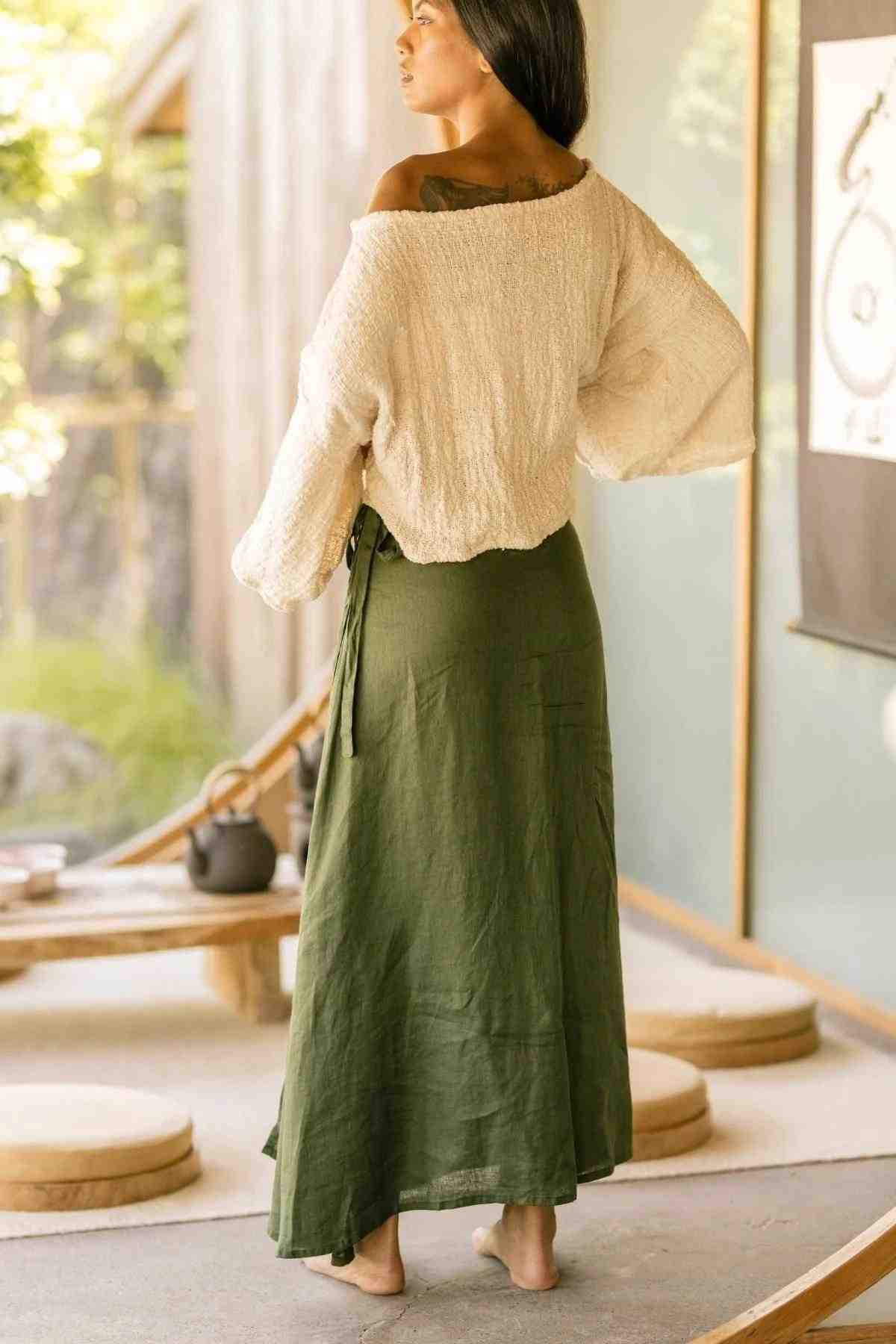 A woman stands barefoot in a sunlit room, wearing a loose white textured off-the-shoulder top and a long Shanti Wrap Skirt by Myrah Penaloza, made of 100% linen in olive green. She is facing away, her head turned slightly to the left as she gazes outside. The room features round floor cushions and minimalistic decor.