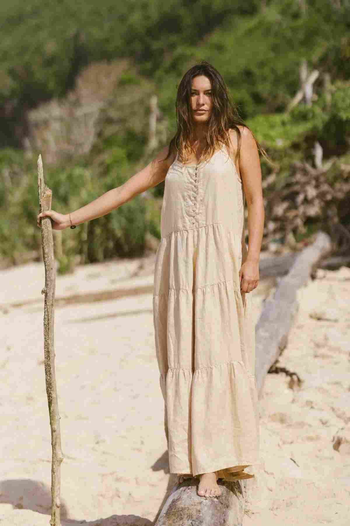 A woman in the La Mexicana Playsuit by Myrah Penaloza, crafted from 100% linen and embodying the essence of resort wear, stands barefoot on a sandy beach. She holds a wooden stick, surrounded by greenery and logs in the background. The sun shines brightly, highlighting her relaxed pose and illuminating her tranquil escape.