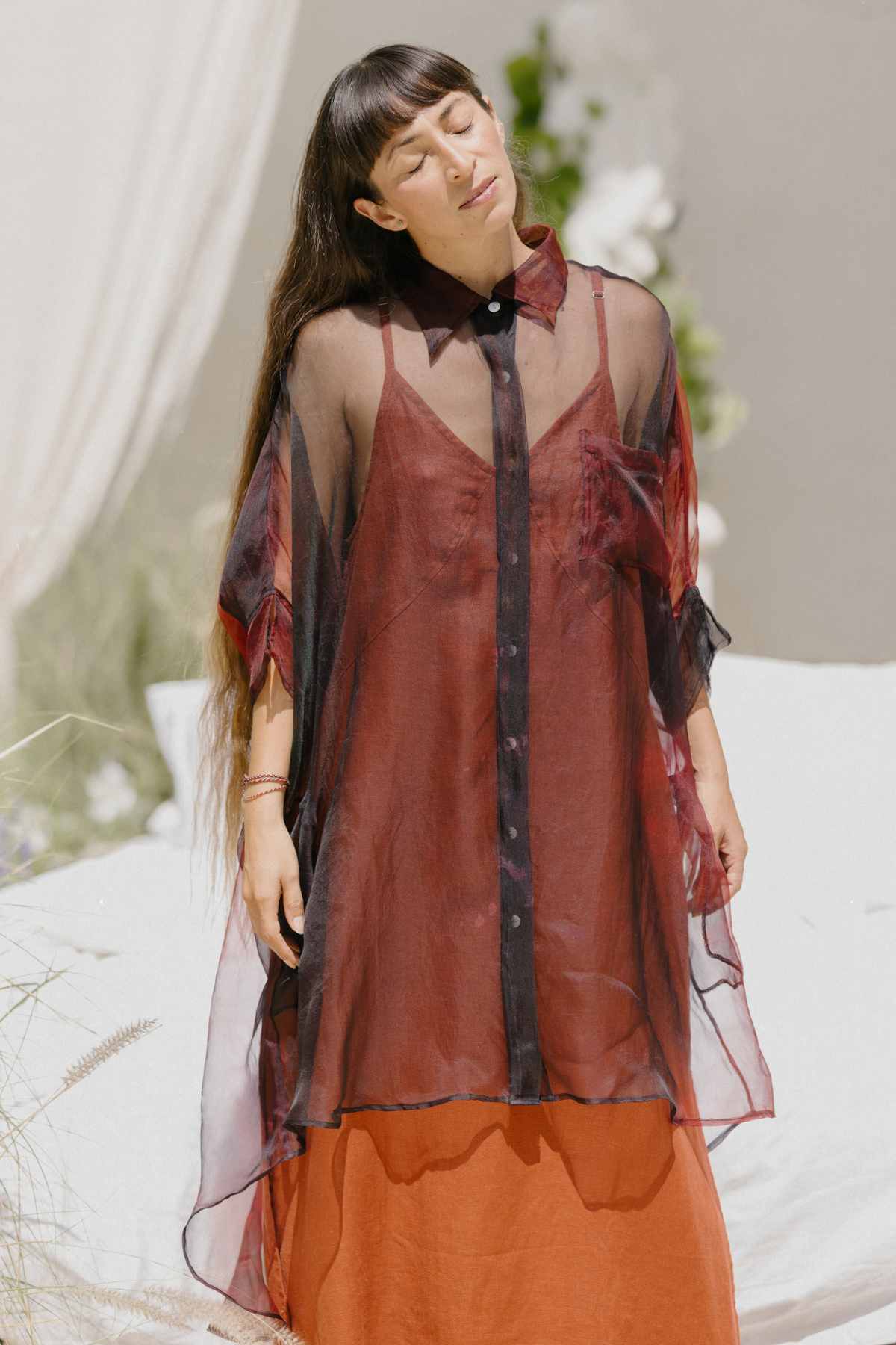 A person with long hair wearing a sheer, red and black Myrah Penaloza Rainbeau Chiffon Silk Suka Top in an oversized fit over a rusty orange dress stands with their eyes closed. White flowers and draped fabric blur in the background, creating a serene and elegant atmosphere.