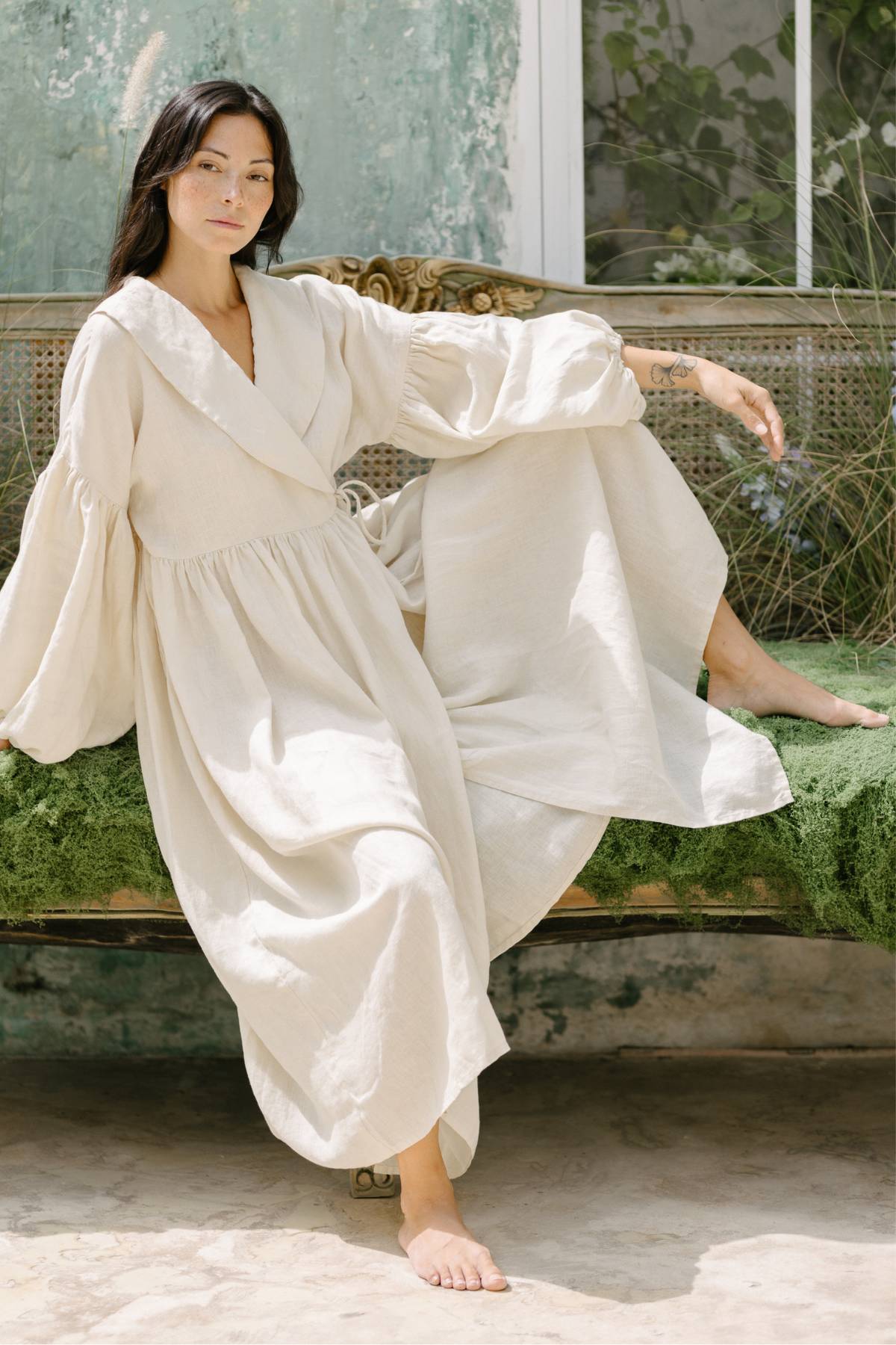 A woman in a flowing Blooming Magdalena Dress by Myrah Penaloza sits barefoot on a green cushioned bench. She is outdoors with a textured wall and plants in the background, exuding a calm and relaxed demeanor.