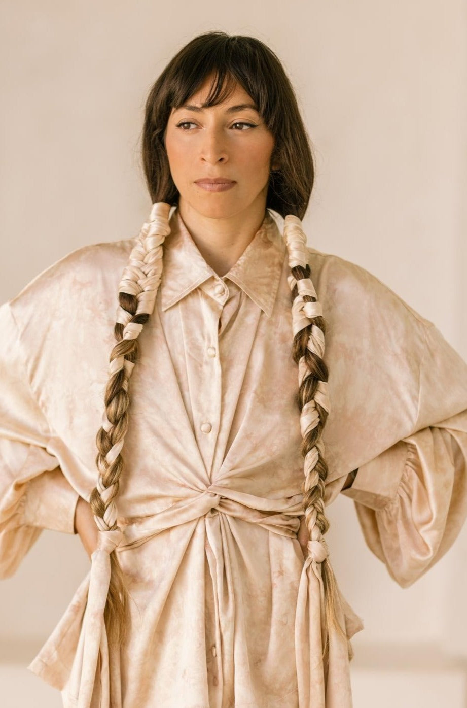 A person with long braided hair, wearing the cream-colored Inanna Top from Myrah Penaloza, crafted from 100% botanically dyed silk, stands slightly turned to the side against a neutral background. The top's loose fit and large sleeves beautifully complement their calm expression.