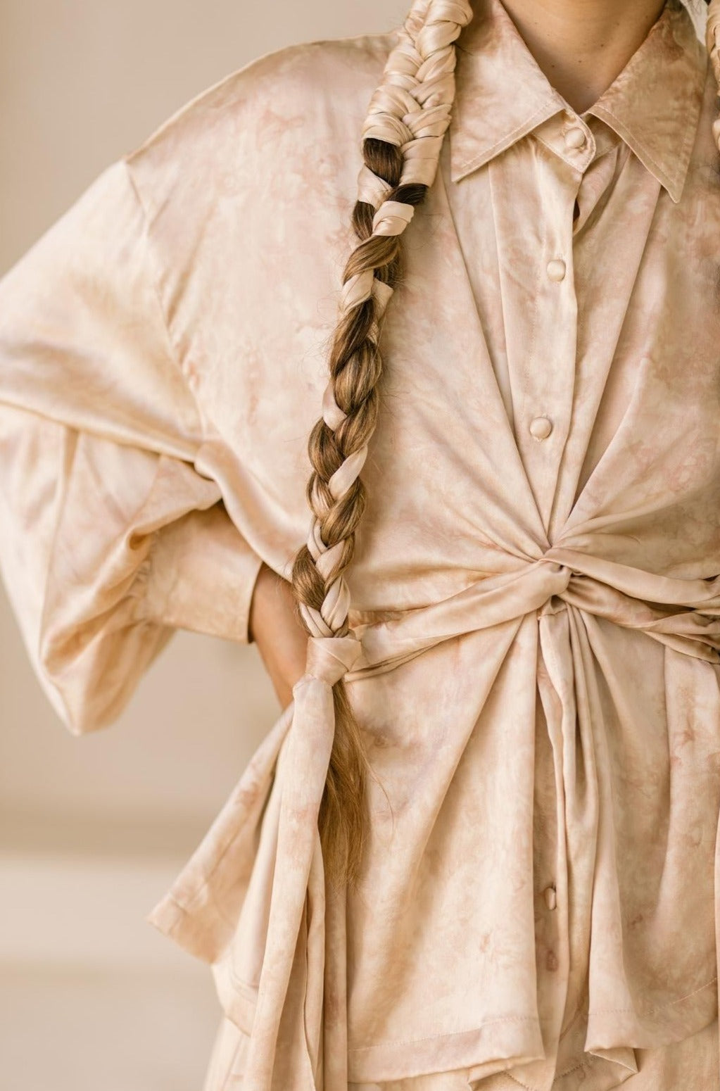 A person wears the Inanna Top Only by Myrah Penaloza, crafted from 100% botanically dyed silk, with a knot tied at the waist. Its beige texture beautifully complements their long hair styled in a braided ponytail draped over the shoulder, reminiscent of an ancient goddess of fertility.