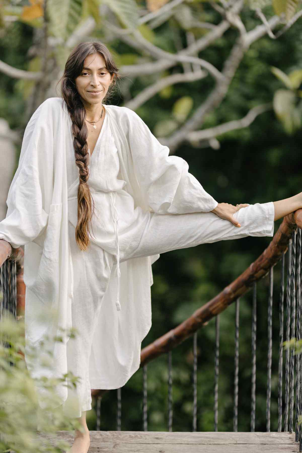 A woman with long dark hair in a braid is dressed in a flowing Myrah Penaloza Dao Robe (100% Linen). She is outdoors on a wooden bridge, stretching one leg onto the railing. The background is lush and green, suggesting a natural setting. She appears calm and relaxed in her attire of natural colors.