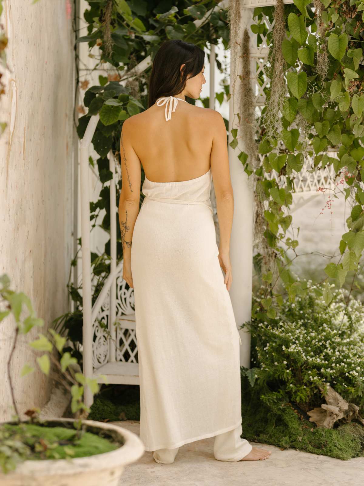 A woman wearing a Honey Cotton Knit Wrap Dress by Myrah Penaloza stands in a lush garden setting. She is facing away from the camera, surrounded by greenery and white garden furniture, with tattoos visible on her arm.