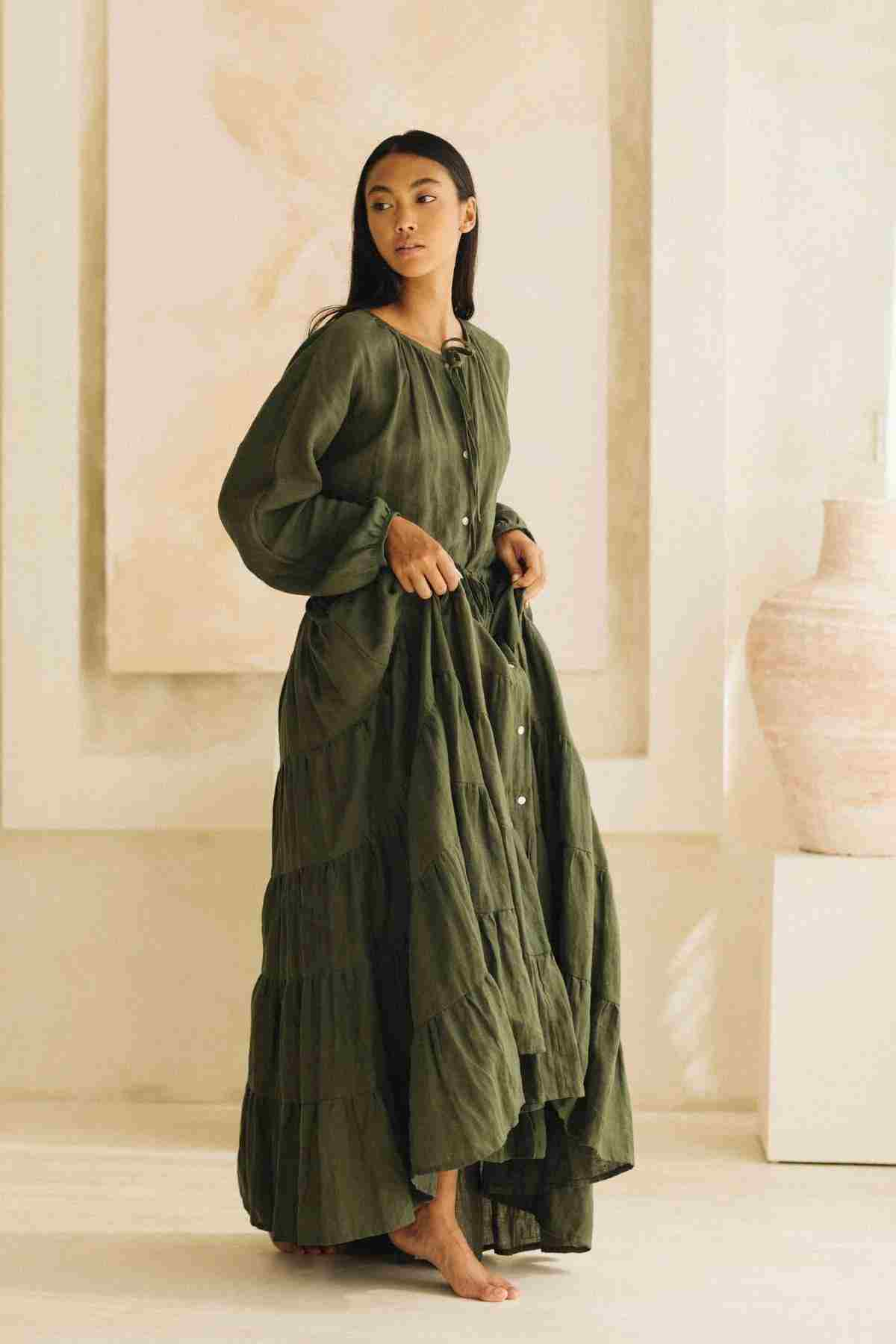 A woman stands barefoot in a light-filled room, wearing the La Mexicana Gown by Myrah Penaloza in a long, flowing olive green linen, adorned with tiered layers and buttons down the front. The gown’s adjustable waist enhances her silhouette as she gazes to the side, with a large vase and neutral-toned artwork complementing the elegant scene.