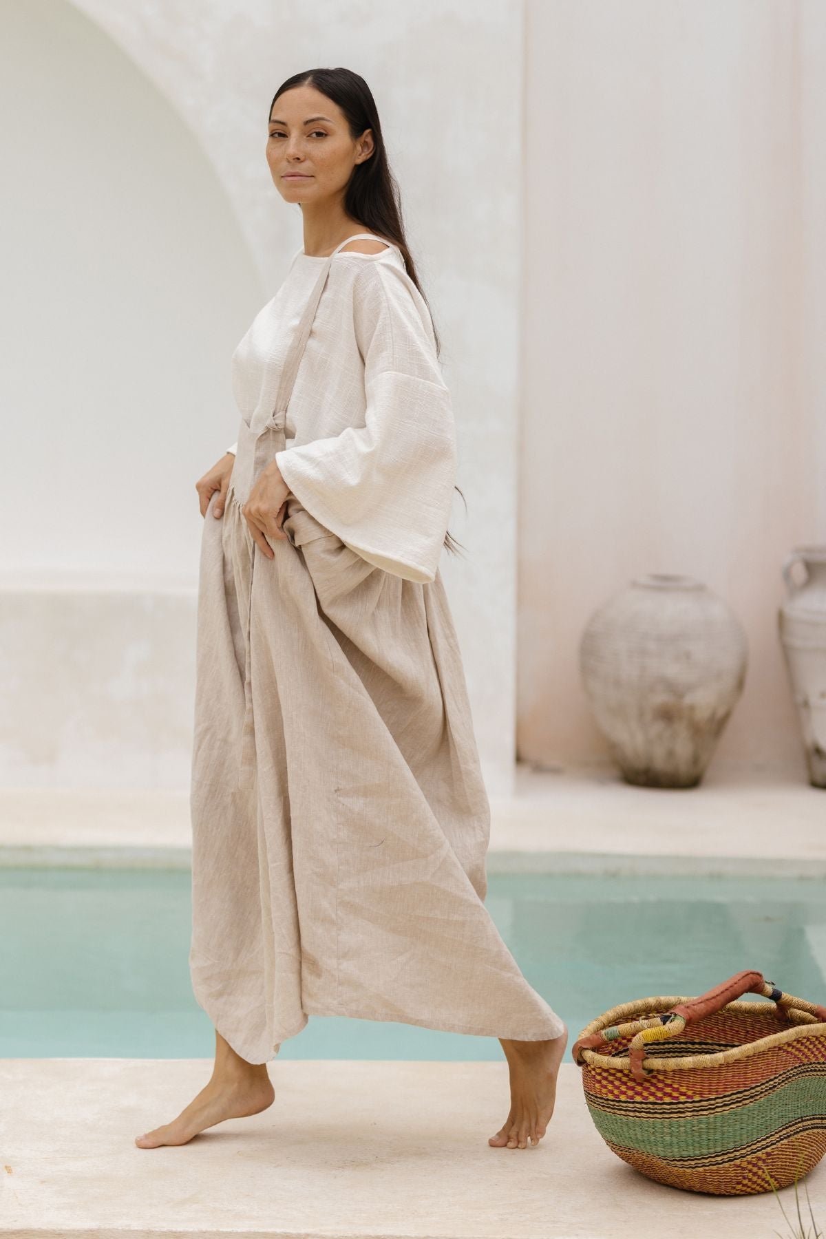 A woman with long dark hair walks barefoot beside a pool, wearing a white long-sleeve top paired with the May May Overall Skirt (Made To Order) by Myrah Penaloza, crafted from beige crinkle linen. In one hand, she carries a small basket adorned with colorful handles. Clay pots are visible in the background, enhancing the serene atmosphere reminiscent of a fairy garden.