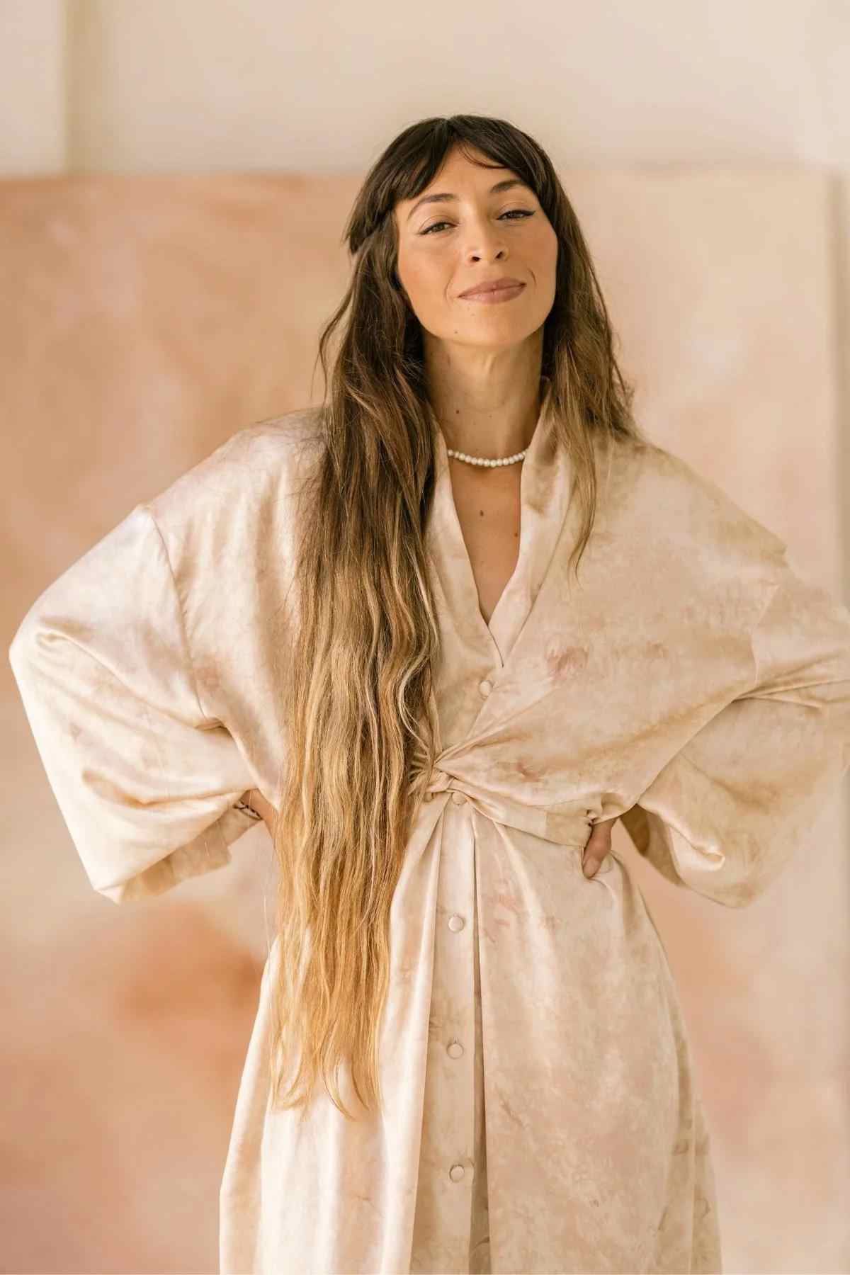 A person with long, wavy hair stands confidently with hands on their hips, wearing a light-colored Myrah Penaloza Silk Inanna Wrap Gown made of botanically hand-dyed silk. They have a calm expression and are set against a soft, blurred background.