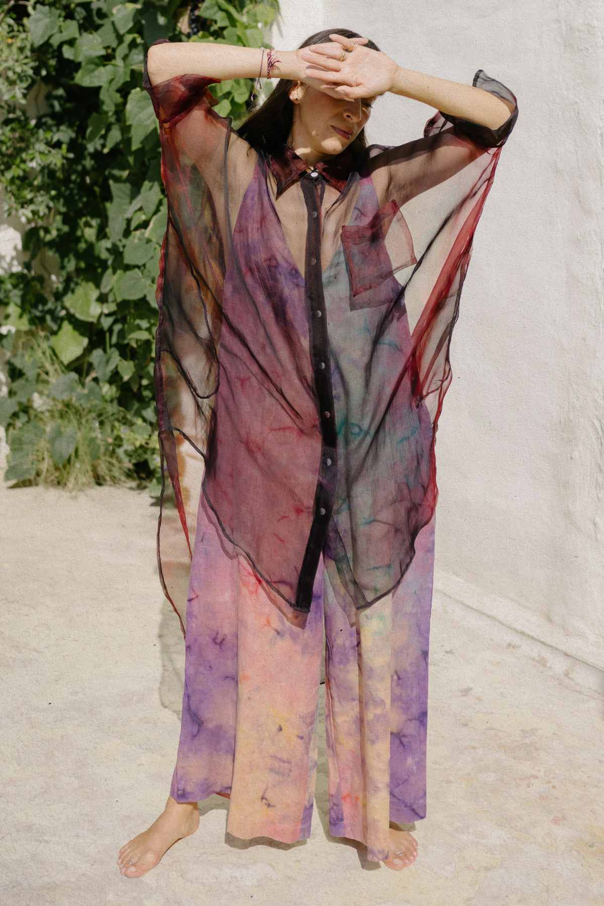 A person stands barefoot outdoors, donning the Myrah Penaloza Rainbeau Chiffon Silk Suka Top over a multi-colored outfit with hues of purple, yellow, and orange. They wear a long, sheer black and red button-up cape. One arm is raised while the other shields their face from the sunlight. Green foliage in the background accentuates their versatile styling options.