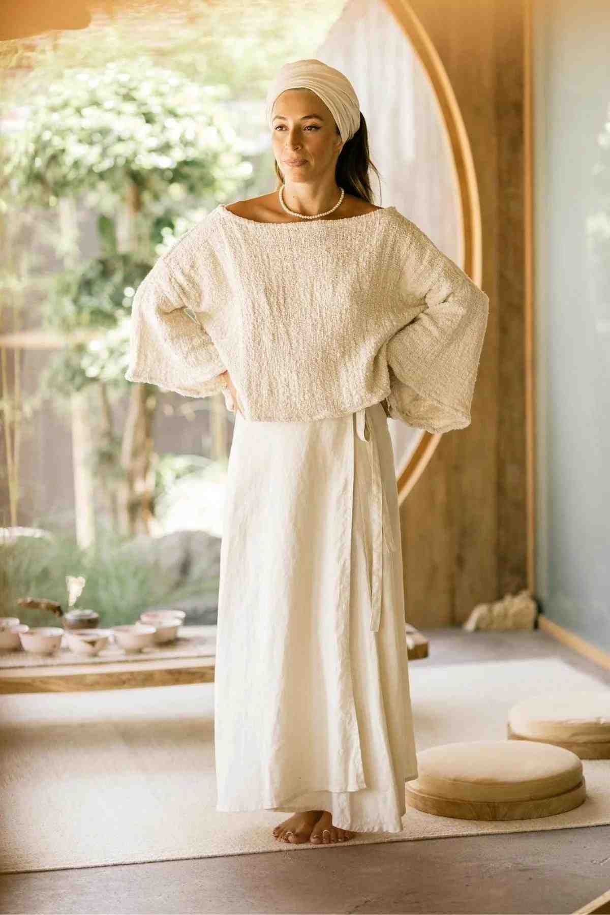 A woman stands indoors in front of a large circular window, wearing a white headscarf and the Spirit Top & Shanti Wrap Skirt Set from Myrah Penaloza. The loose-fitting cream-colored top is made from hand-woven Tummangal cotton and is paired with an adjustable cream skirt. The room exudes a serene ambiance with soft natural light, minimalist decor, and seating cushions.