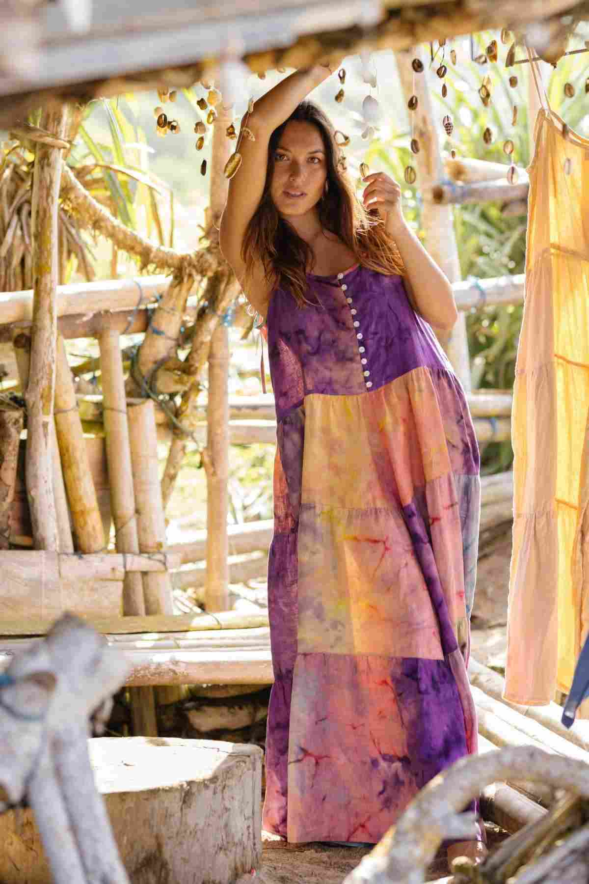 A woman wearing the La Mexicana Playsuit by Myrah Penaloza stands in a rustic wooden structure adorned with hanging shells. She lifts her arm to touch the frame and gazes at the camera, as sunlight filters through, creating a warm, natural atmosphere.